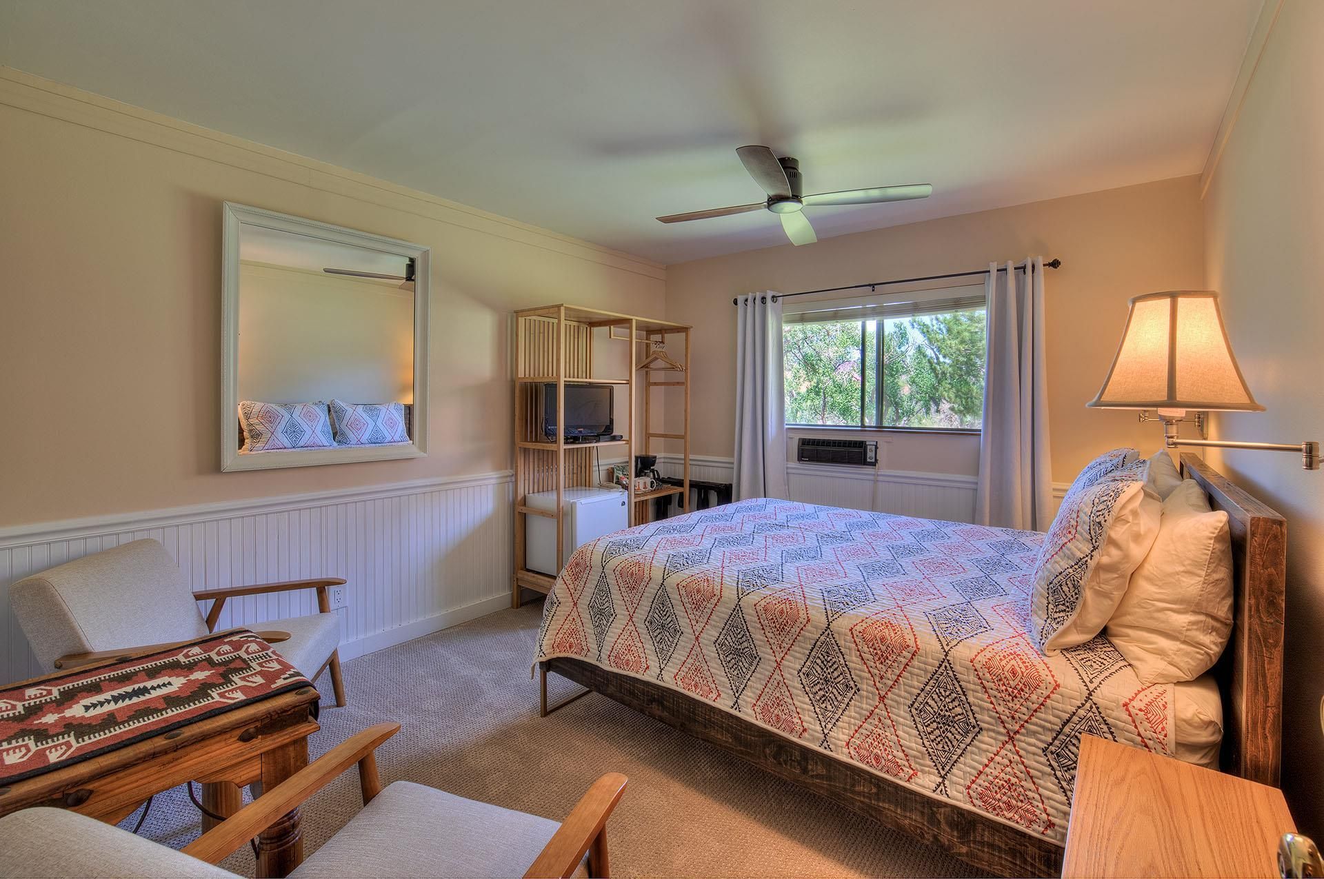 Bright bedroom with patterned bedspread, window, lamps, and upholstered chair by a wall mirror