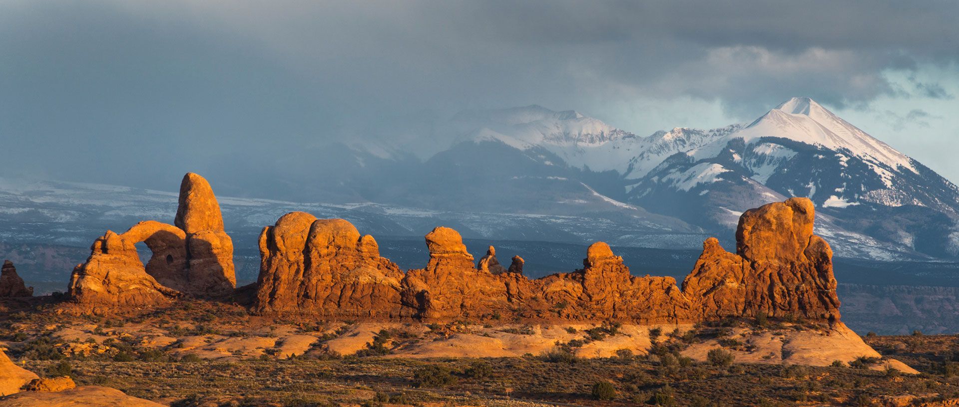 Orange sandstone formations at sunset with snowcapped mountains and cloudy sky in the background