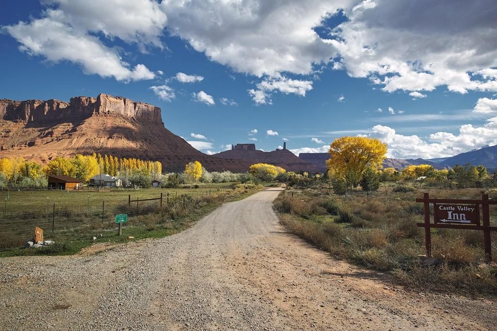 Gravel road leading through autumn fields toward red cliffs under a blue sky with clouds