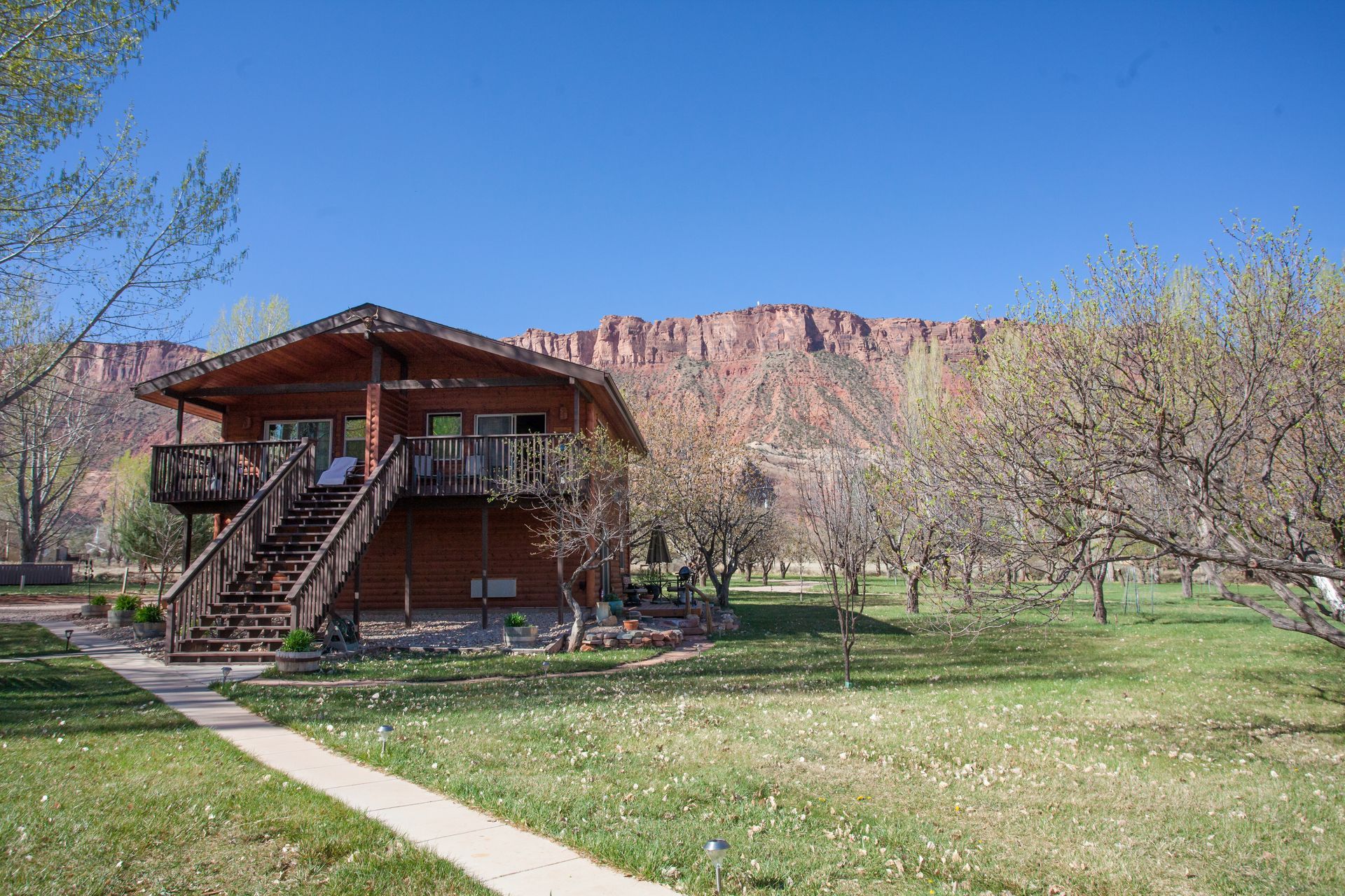 Rustic cabin in a grassy orchard beneath red cliffs under a clear blue sky