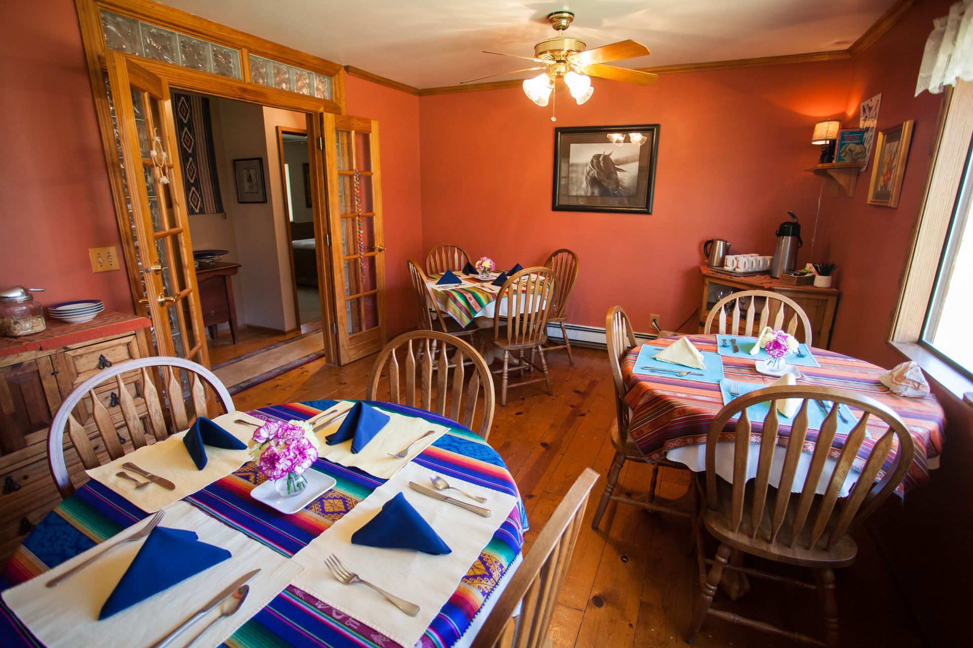 Dining room with several set tables, wooden chairs, and warm orange walls under a ceiling fan.