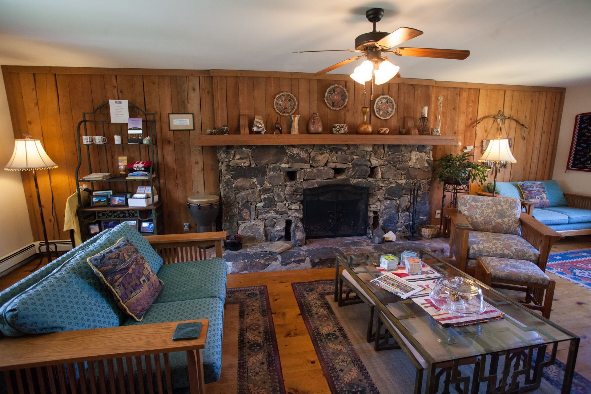 Cozy living room with stone fireplace, wood-paneled walls, ceiling fan, and blue seating area