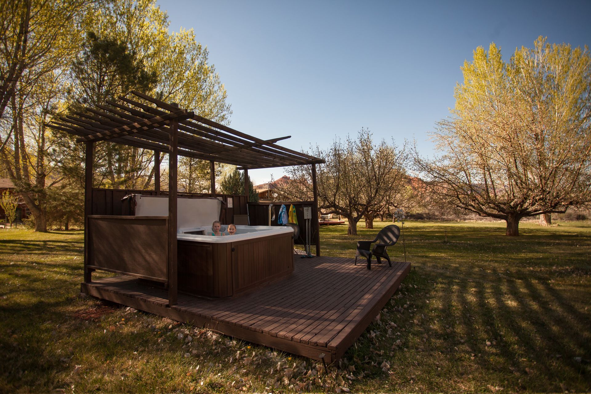 Rustic outdoor kitchen pergola on a grassy lawn with trees and benches in warm sunlight