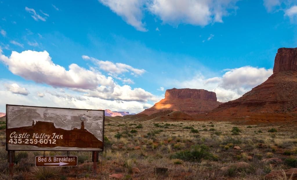 Desert landscape with red rock mesas under a blue sky and a roadside sign in the foreground.