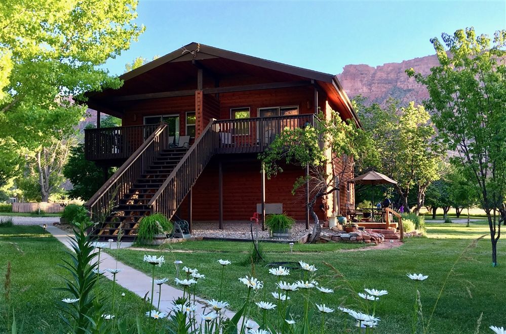 Rustic cabin with front porch and stairs, surrounded by green lawn and trees in bright daylight