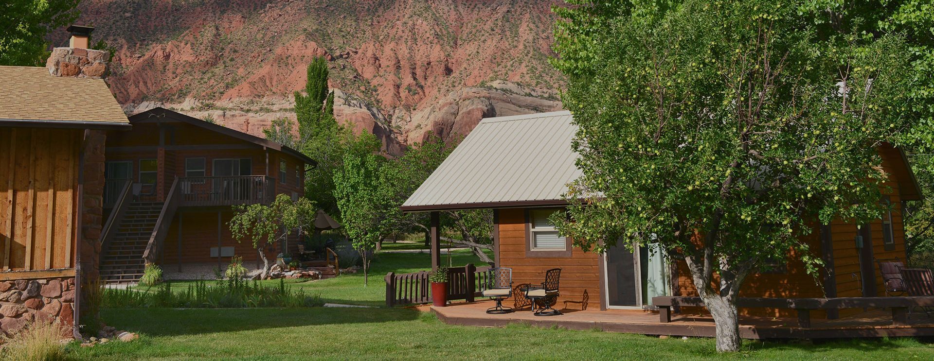 Rustic cabins on a green lawn beside a rocky cliff, with trees and patio chairs in warm sunlight