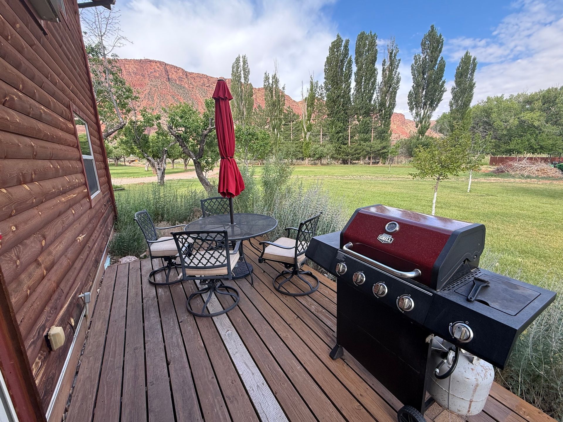 Patio deck with outdoor dining table, red umbrella, and black gas grill beside a grassy yard and hills.