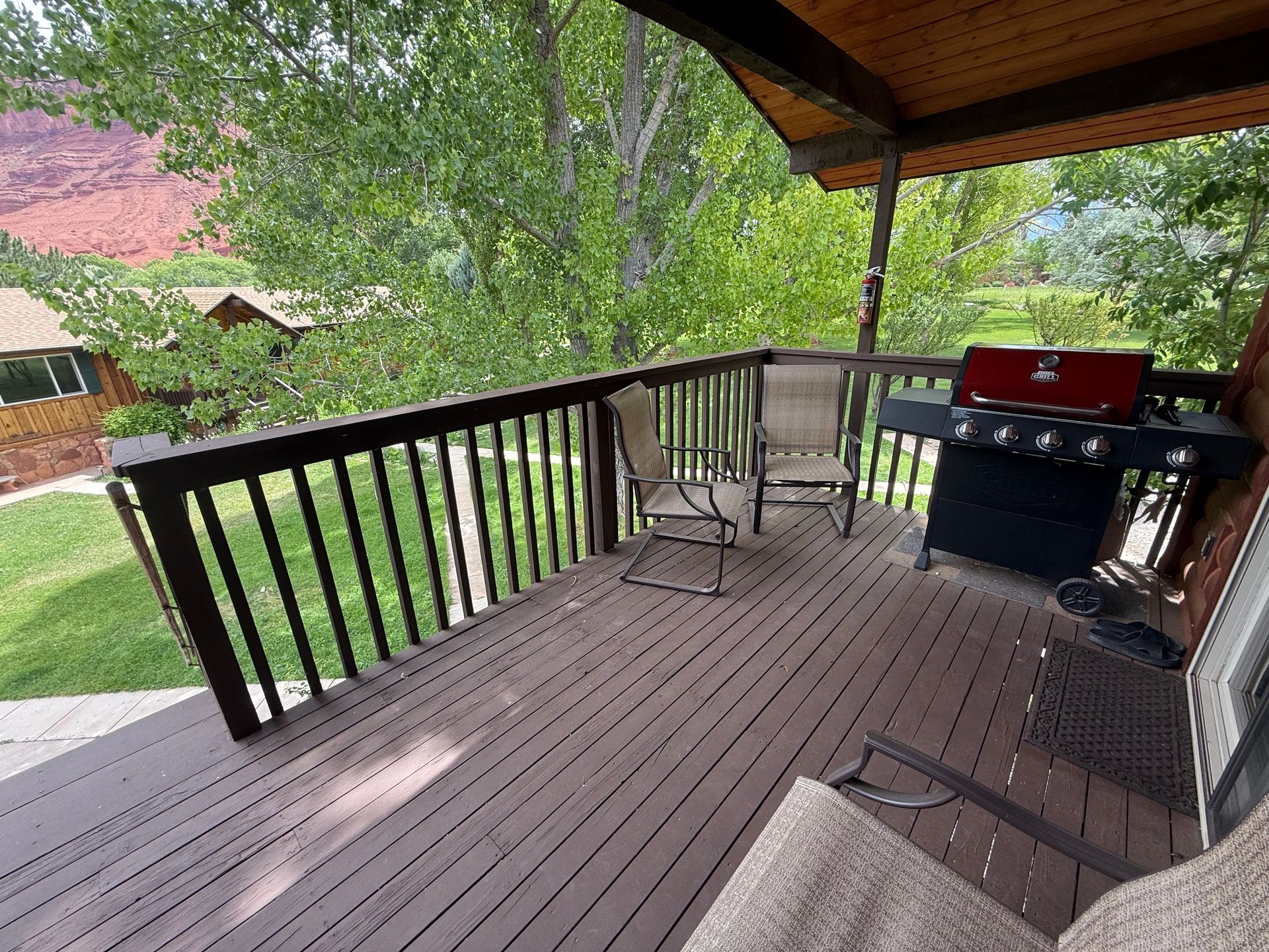 Covered wooden deck with black railing, grill, and patio chairs overlooking a green yard and mountains.