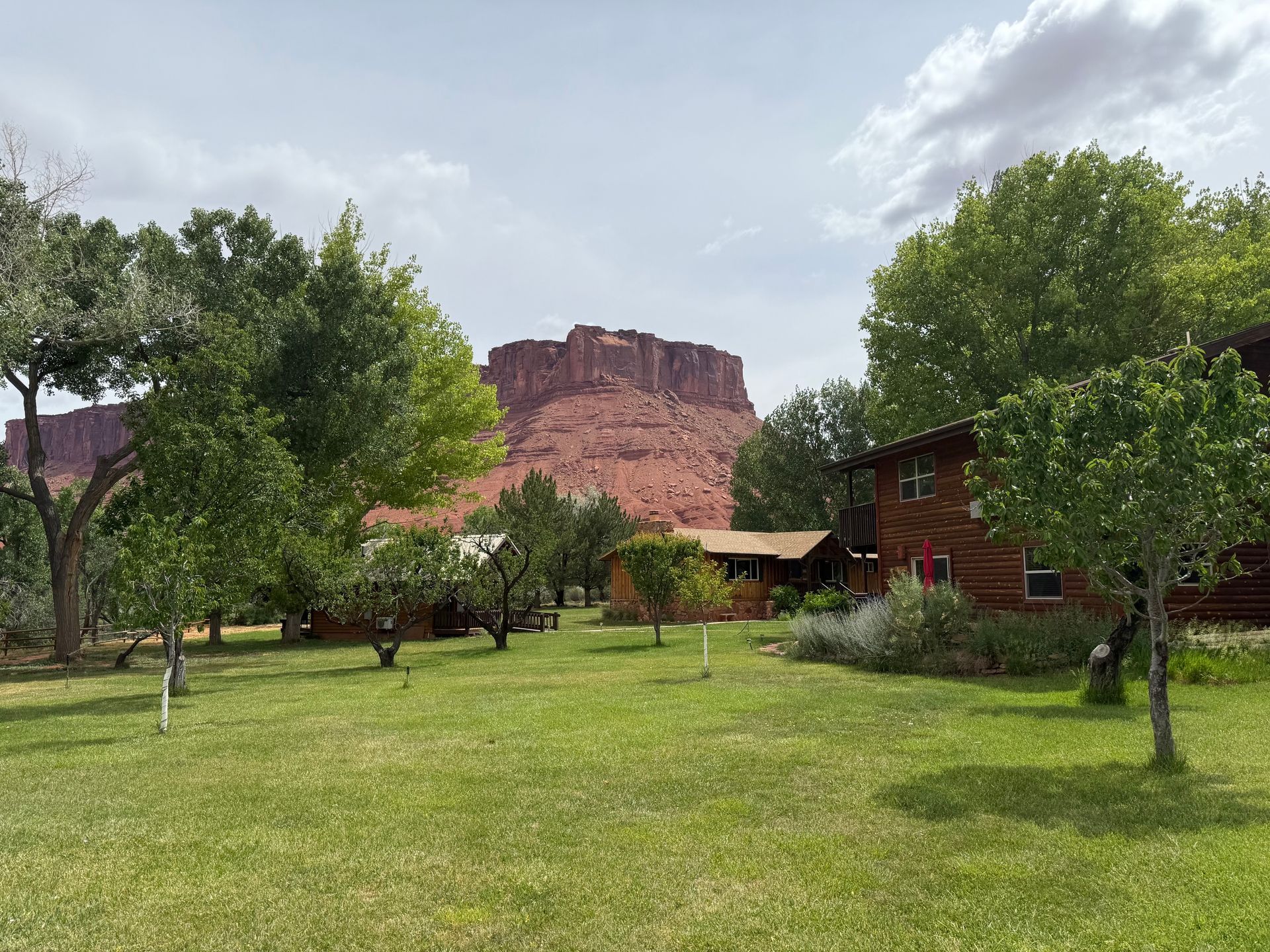 Grassy park with trees and cabins in front of a red sandstone mesa under a cloudy sky