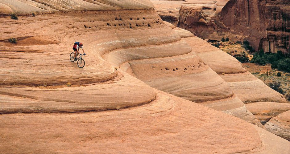 Mountain biker riding down a tan sandstone desert slope with layered rock formations.