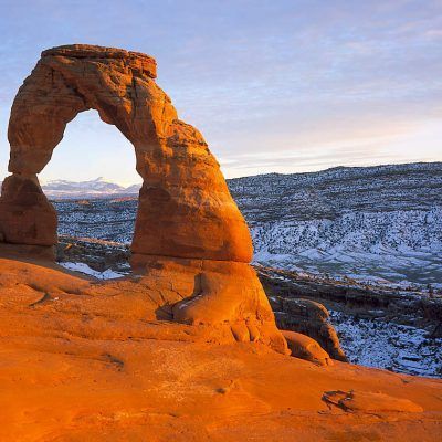 Snow-dusted desert arch at sunset with warm orange sandstone and distant mountains