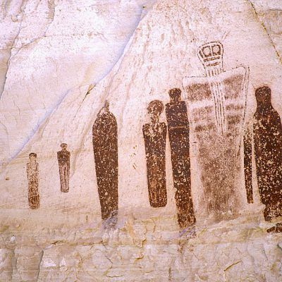 Ancient cave painting of several tall figures on a rock wall, in dark brown and white.