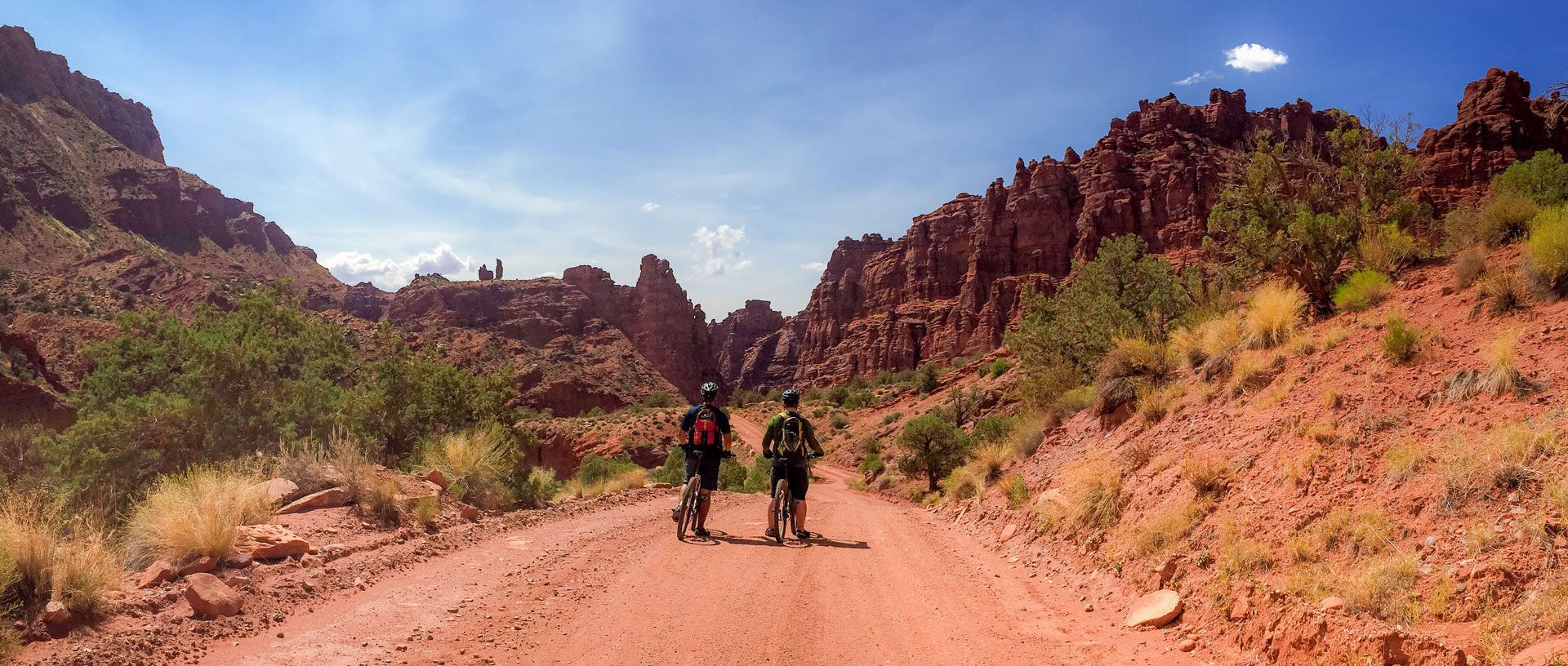 Two hikers on a dusty desert trail between red rock cliffs under a blue sky