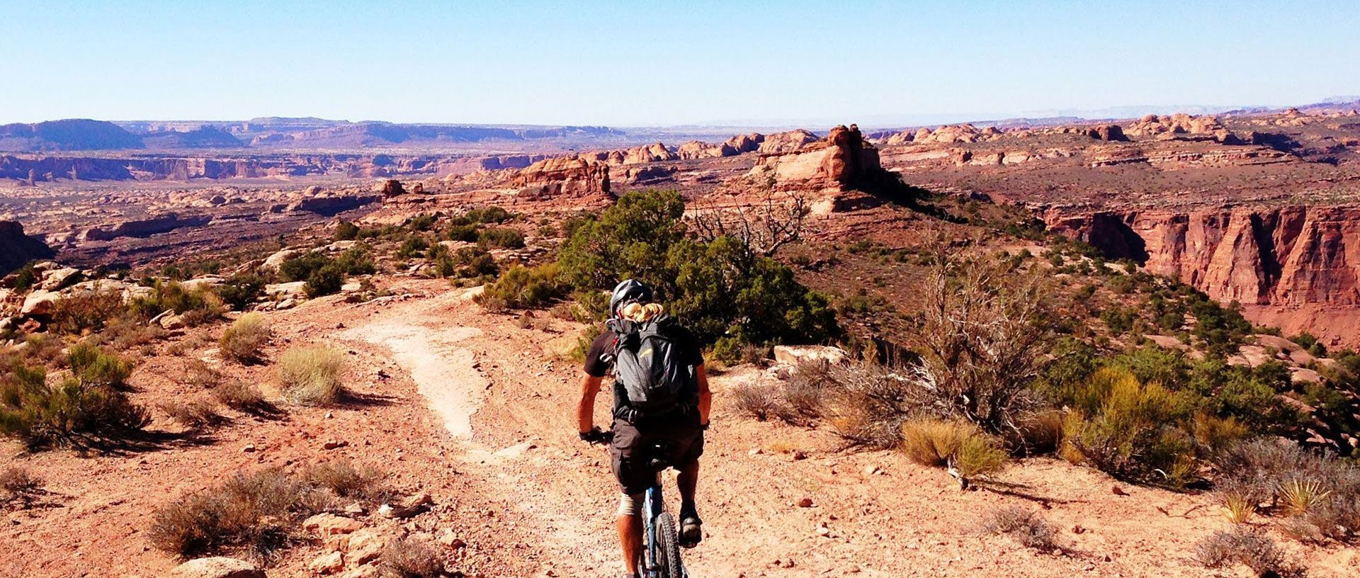 Hiker walking on a dusty desert trail with canyon cliffs and distant mountains under a clear sky