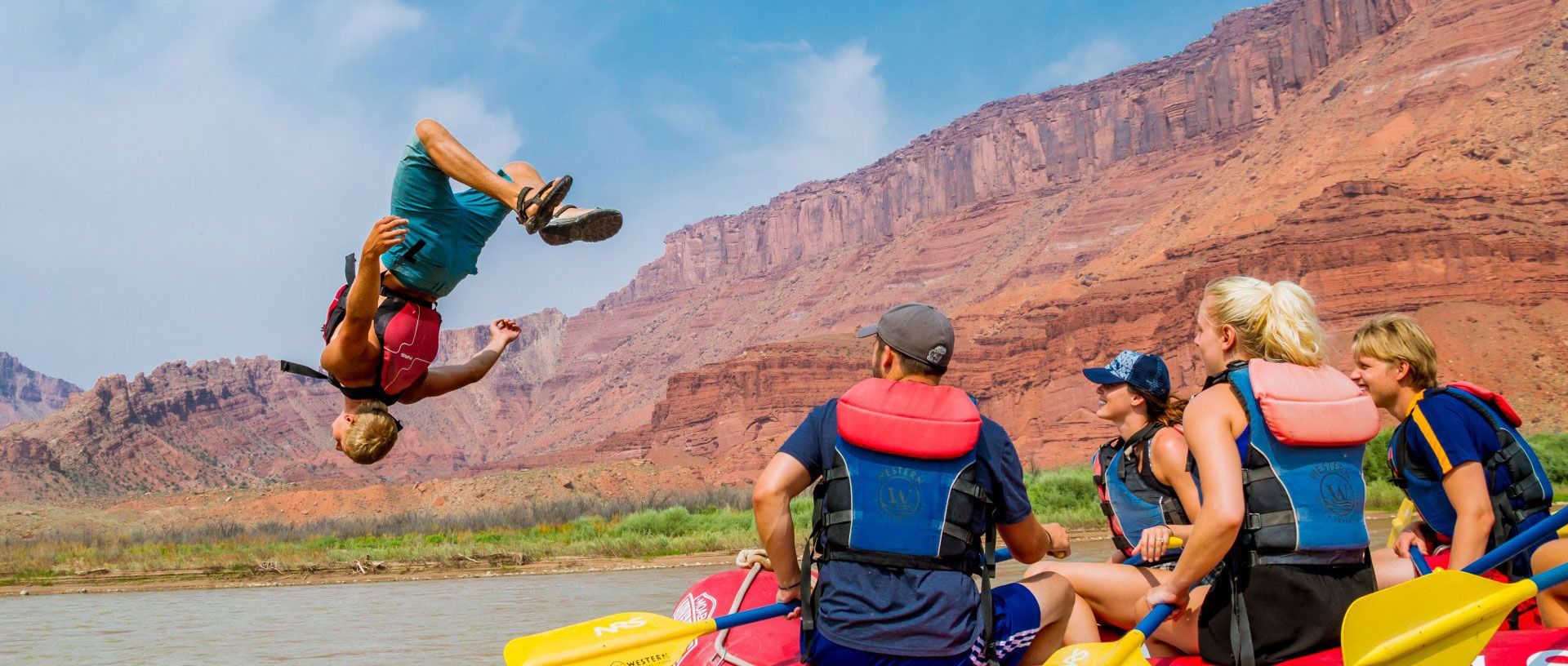 Rafters watch as a person flips backward into a red kayak on a desert river with red cliffs