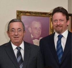 Two men in suits standing indoors before a framed portrait