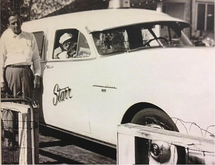 Two people beside a white car with a man in the driver's seat at a wooden gate