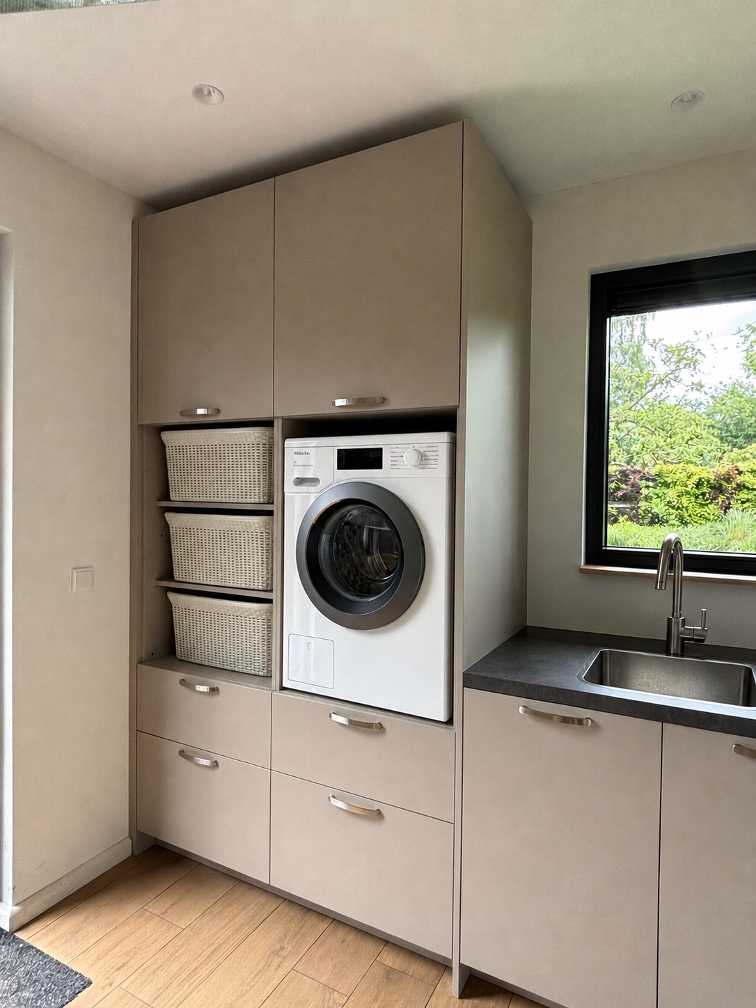 Compacte wasruimte met beige kasten, een wasmachine met voorlader en een kleine wasbak bij een raam.