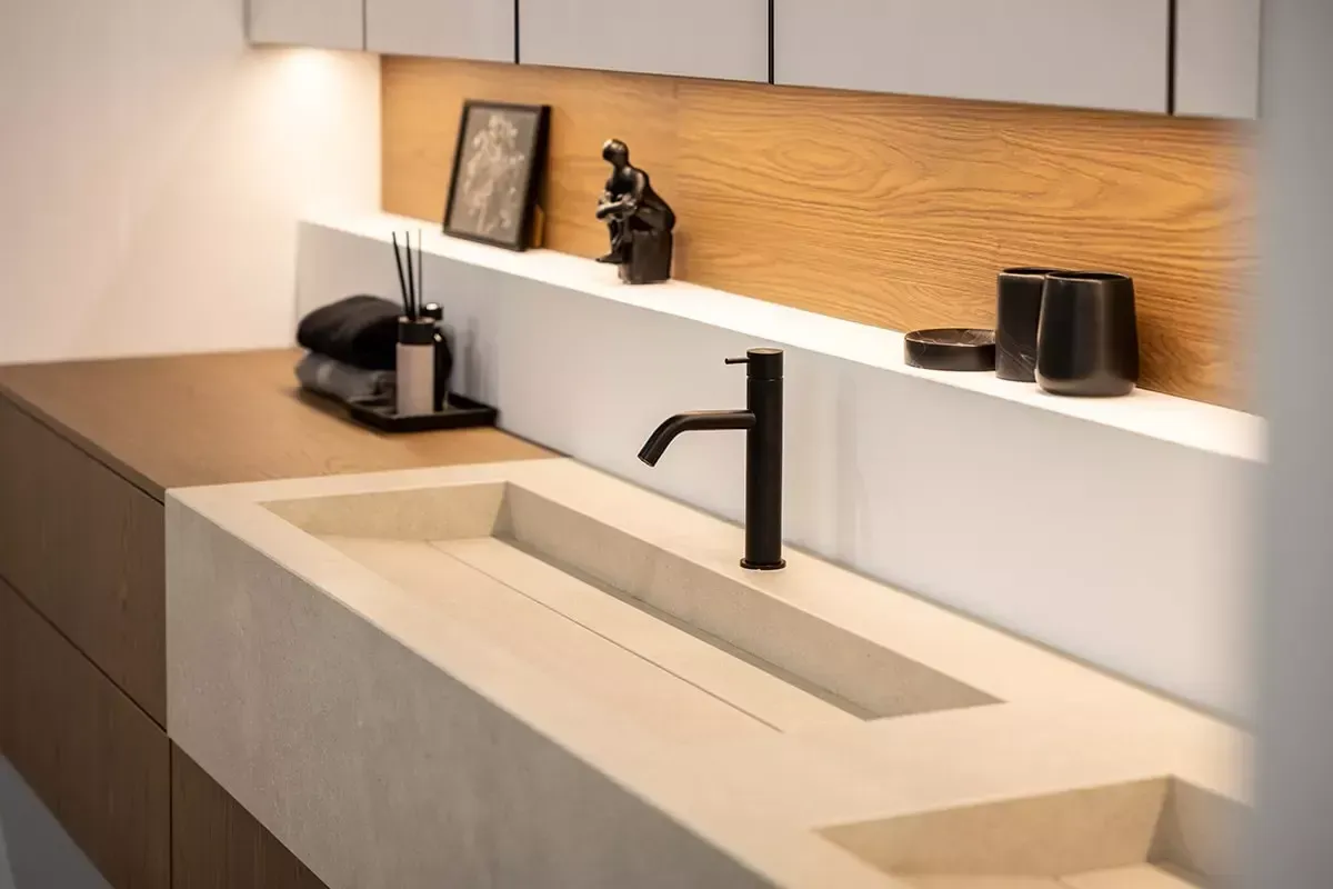 Moderne, minimalistische badkamerkast met betonnen wastafel, zwarte kraan, houten plank en kleine decoratieve items.
