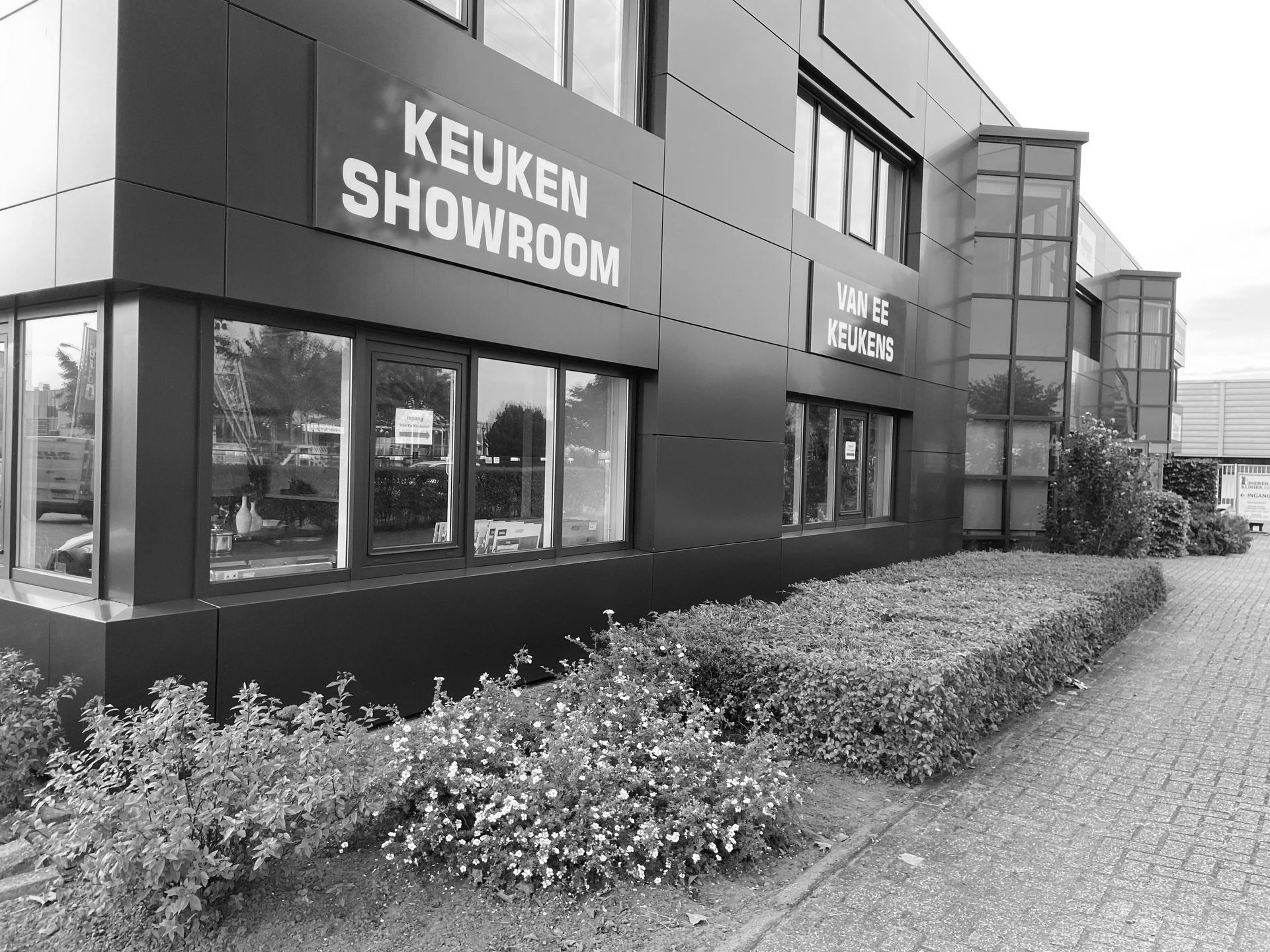 Zwart-wit exterieur van een keukenshowroom met grote ramen en hagen langs de stoep.