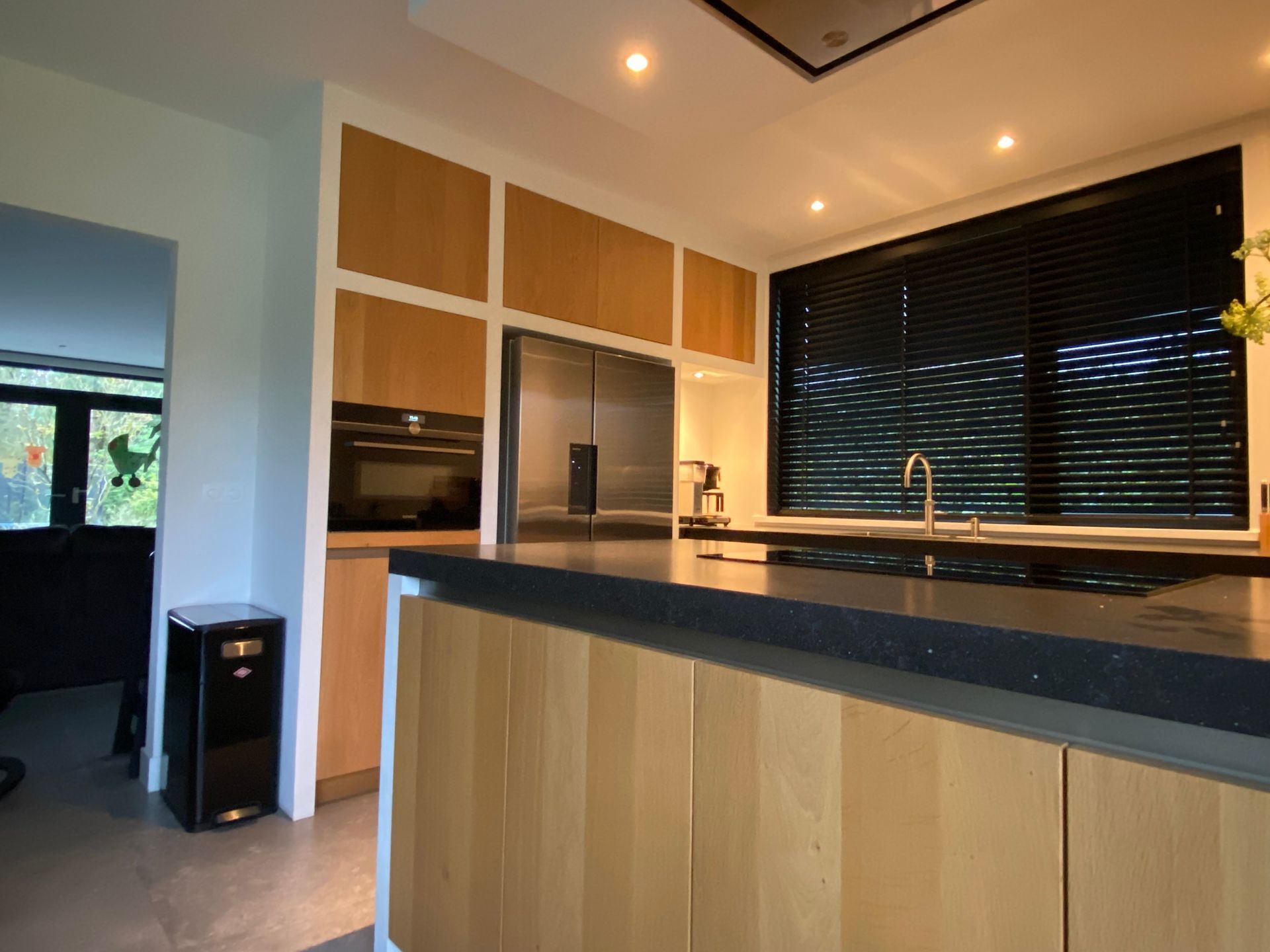 Moderne keuken met houten kasten, zwarte werkbladen en grote raamjaloezieën.
