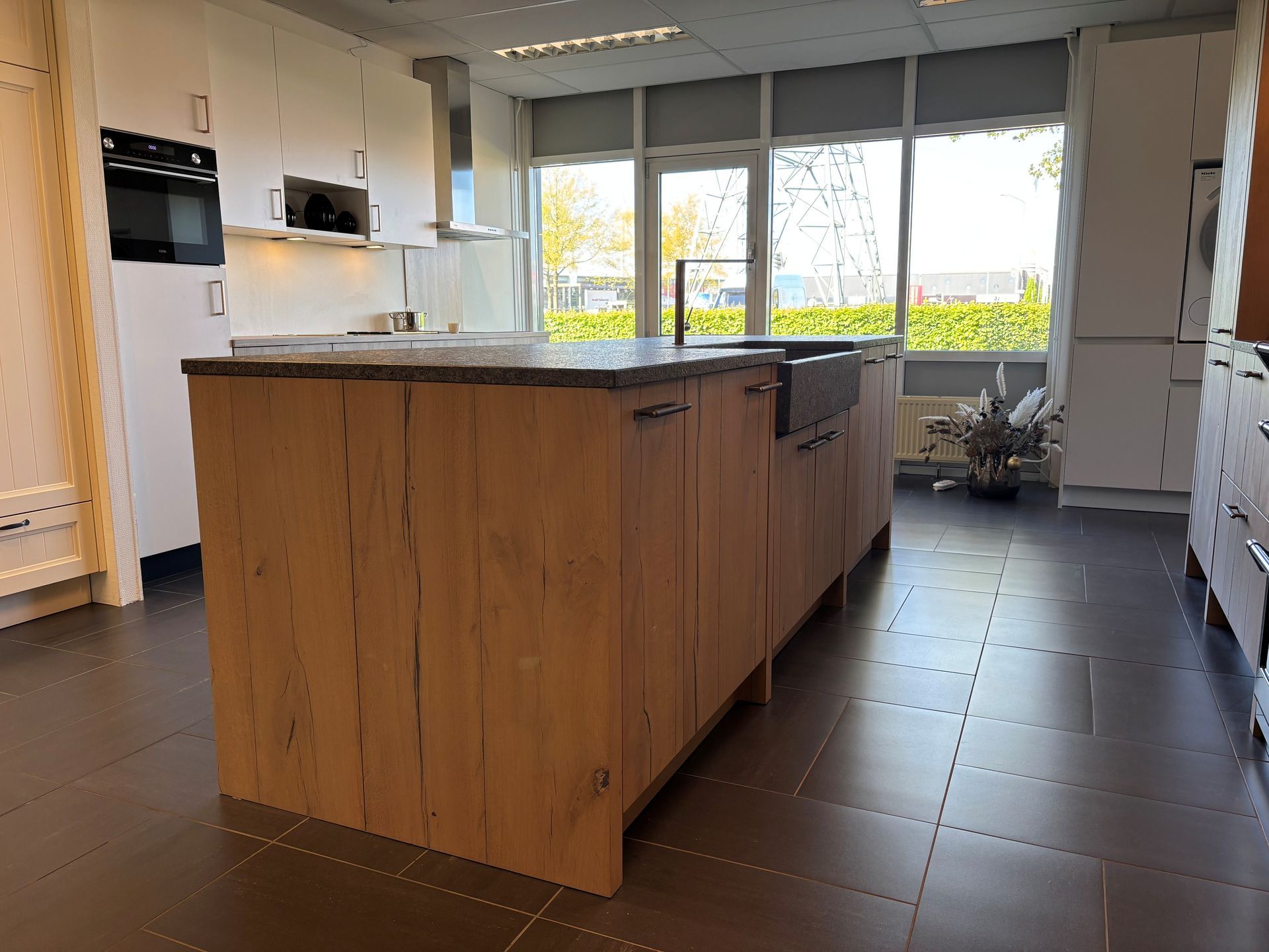 Moderne keuken met een groot houten kookeiland, witte kasten, een donkere tegelvloer en ramen met uitzicht op groen.