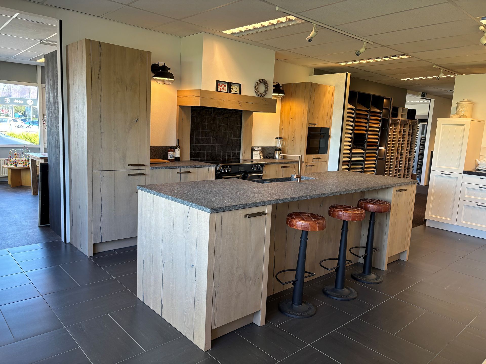Moderne keukenshowroom met houten kasten, grijs kookeiland en drie rode barkrukken.