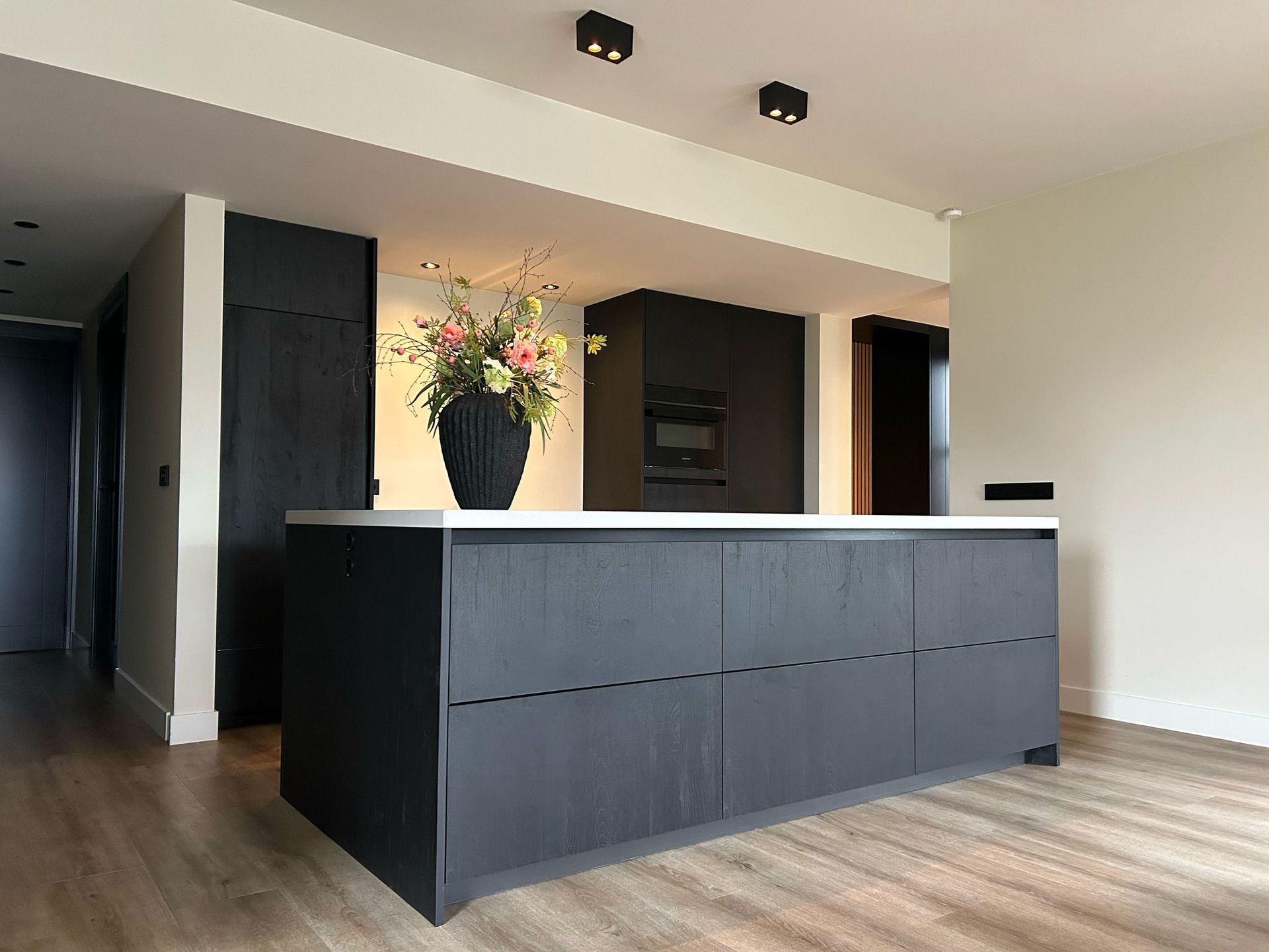 Moderne keuken met groot donker kookeiland, witte werkbladen en een vaas met bloemen in een lichte, open ruimte.