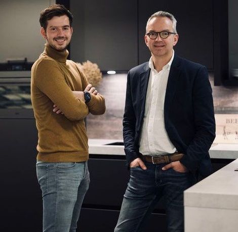 Twee mannen staan in een moderne keuken, de ene glimlachend met de armen over elkaar, de andere in een blazer en spijkerbroek.