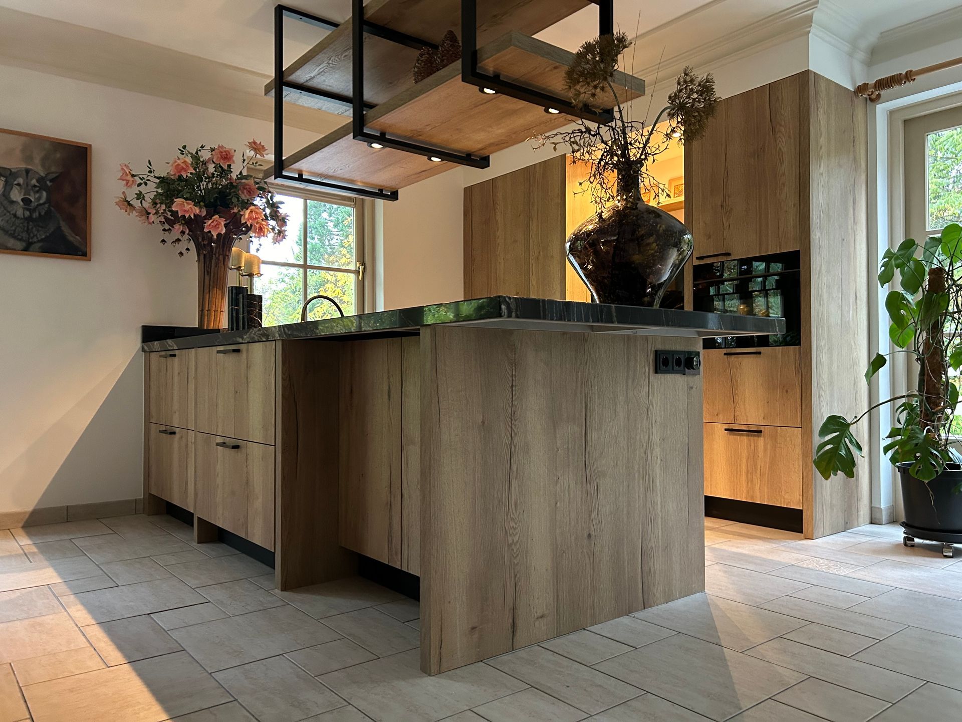 Moderne keuken met houten kookeiland, zwarte werkbladen, hanglamp en lichte ramen.