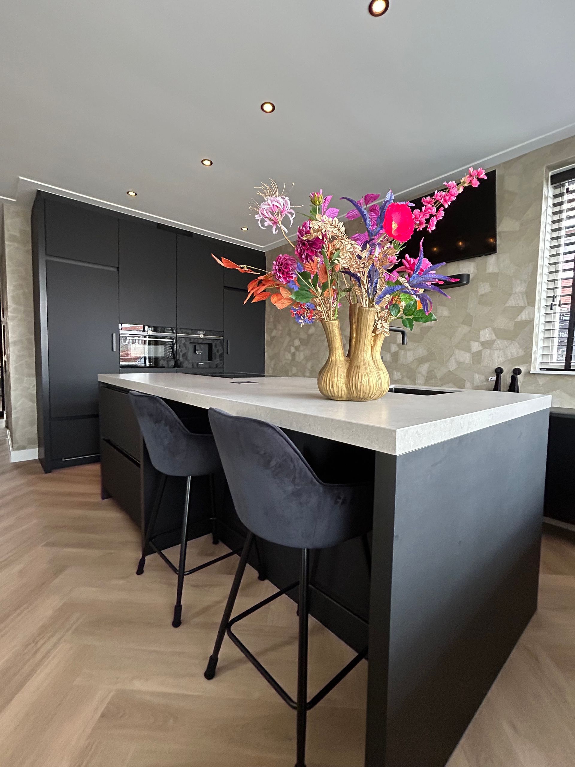 Moderne keuken met zwart kookeiland, twee barkrukken en een vaas met kleurrijke bloemen.