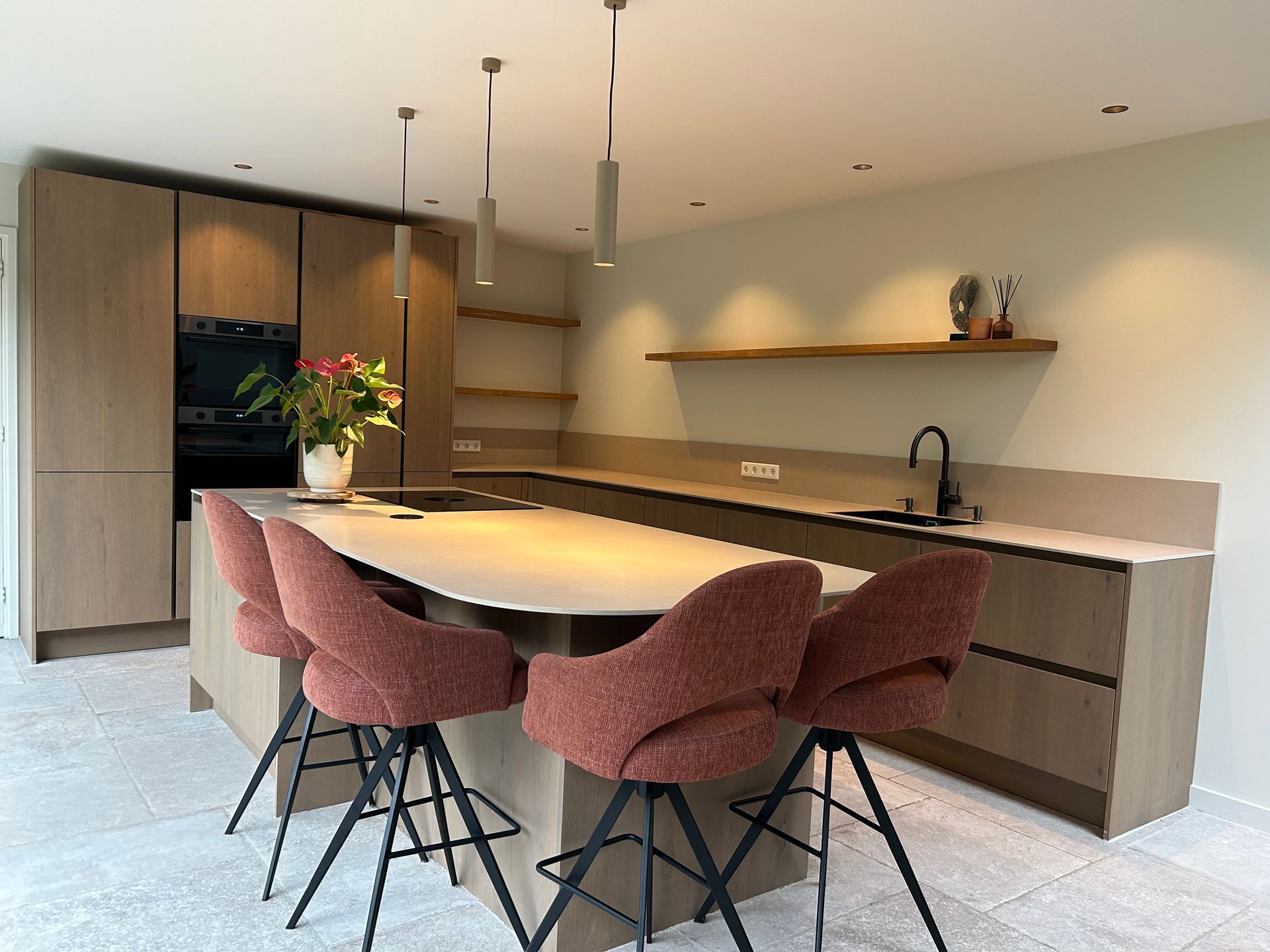 Moderne keuken met houten kasten, een gebogen kookeiland, roze krukken en minimalistische planken aan beige muren.