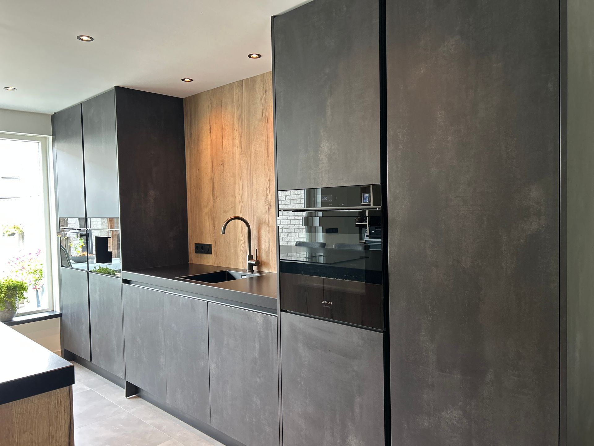 Moderne, minimalistische keuken met donkere kasten, houten lambrisering en een zwart aanrechtblad, gelegen in een lichte hal.