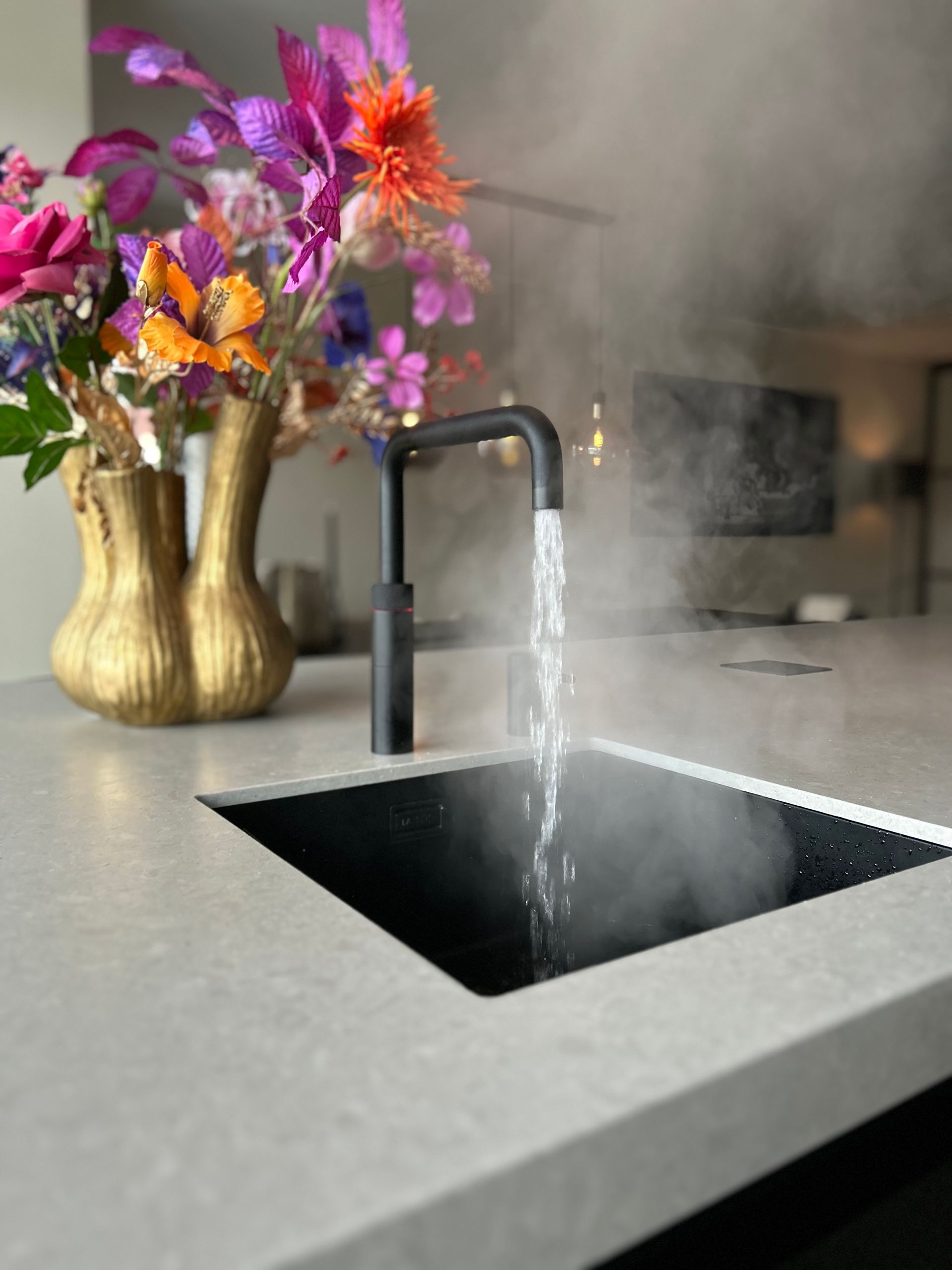 Keukenwastafel met stromend water, zwarte kraan en een gouden vaas met kleurrijke bloemen op een grijs aanrechtblad.