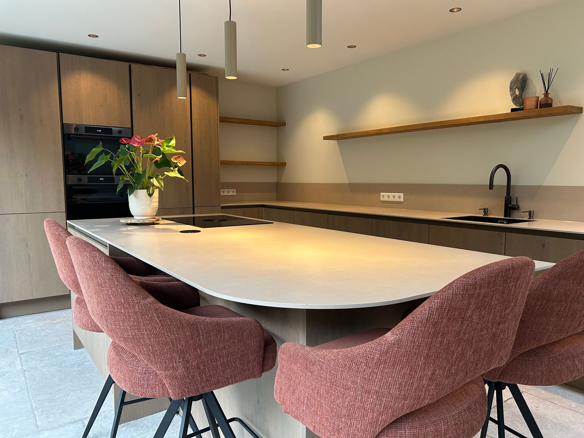 Moderne keuken met een groot kookeiland, roze stoelen, houten kasten en een vaas met bloemen.