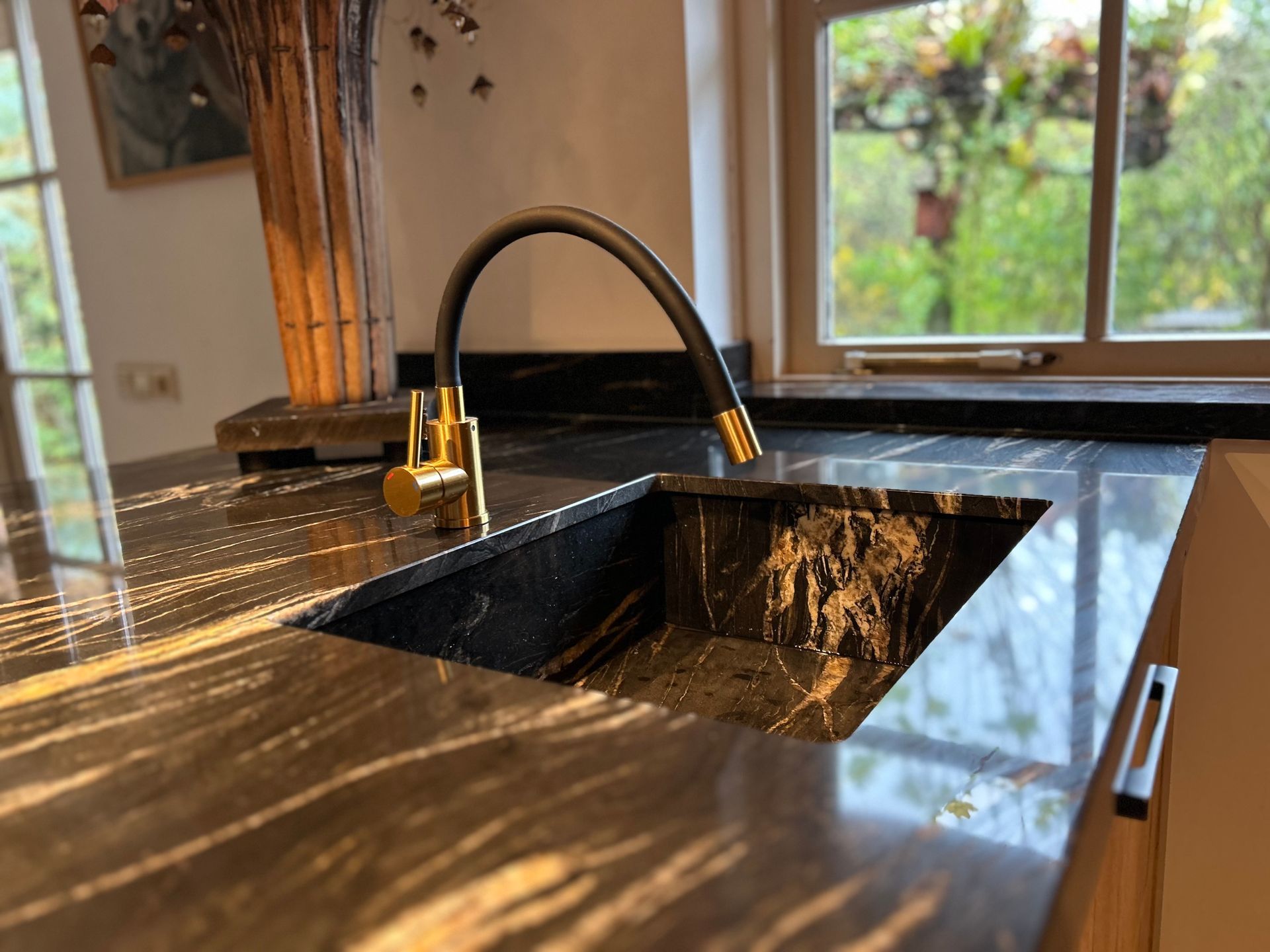 Moderne keuken met zwart marmeren aanrechtblad, matzwarte spoelbak en gouden kraan bij een raam.