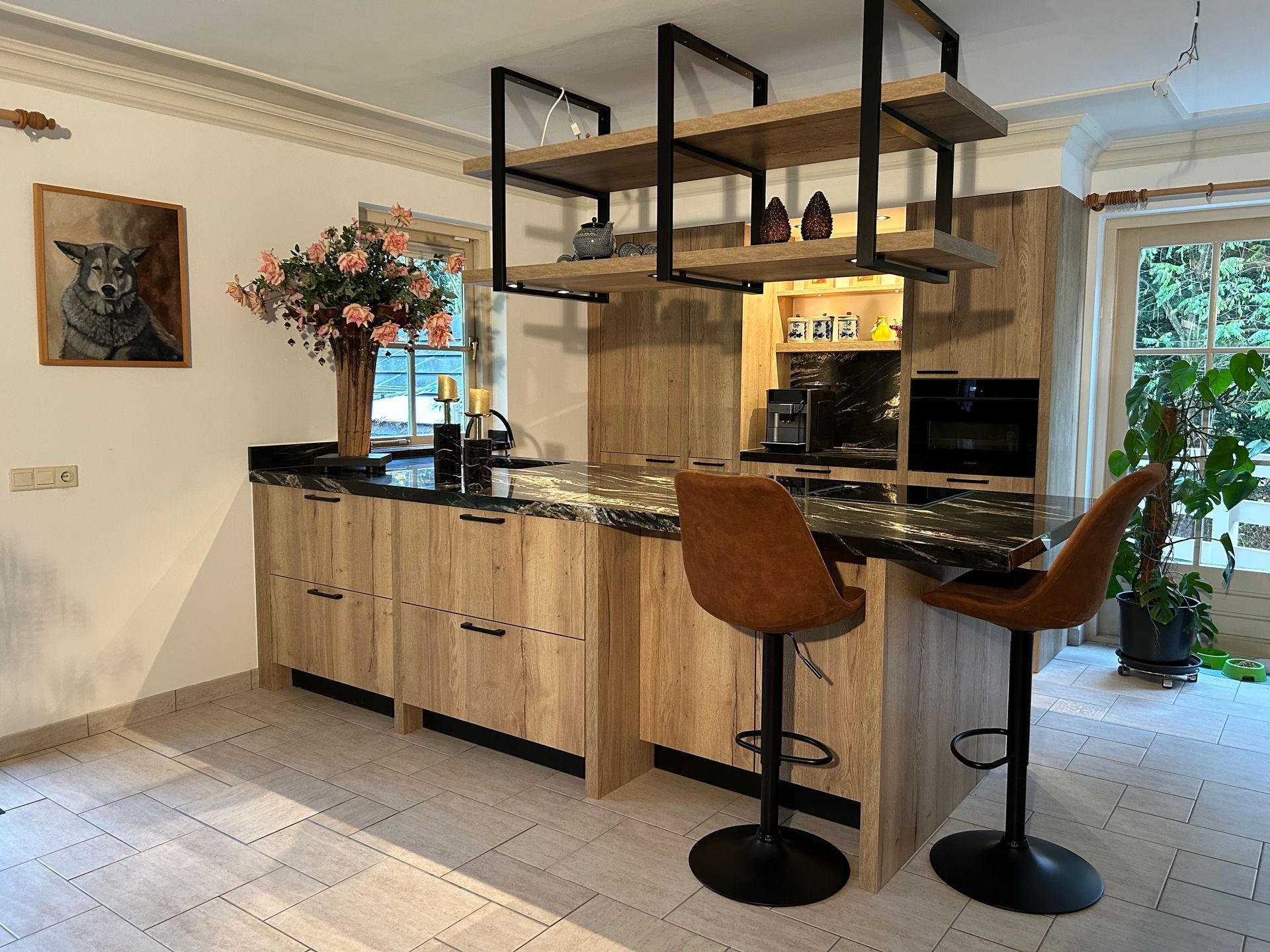 Moderne keuken met lichte houten kasten, zwart aanrechtblad, barkrukken aan het kookeiland en open planken bij een raam.