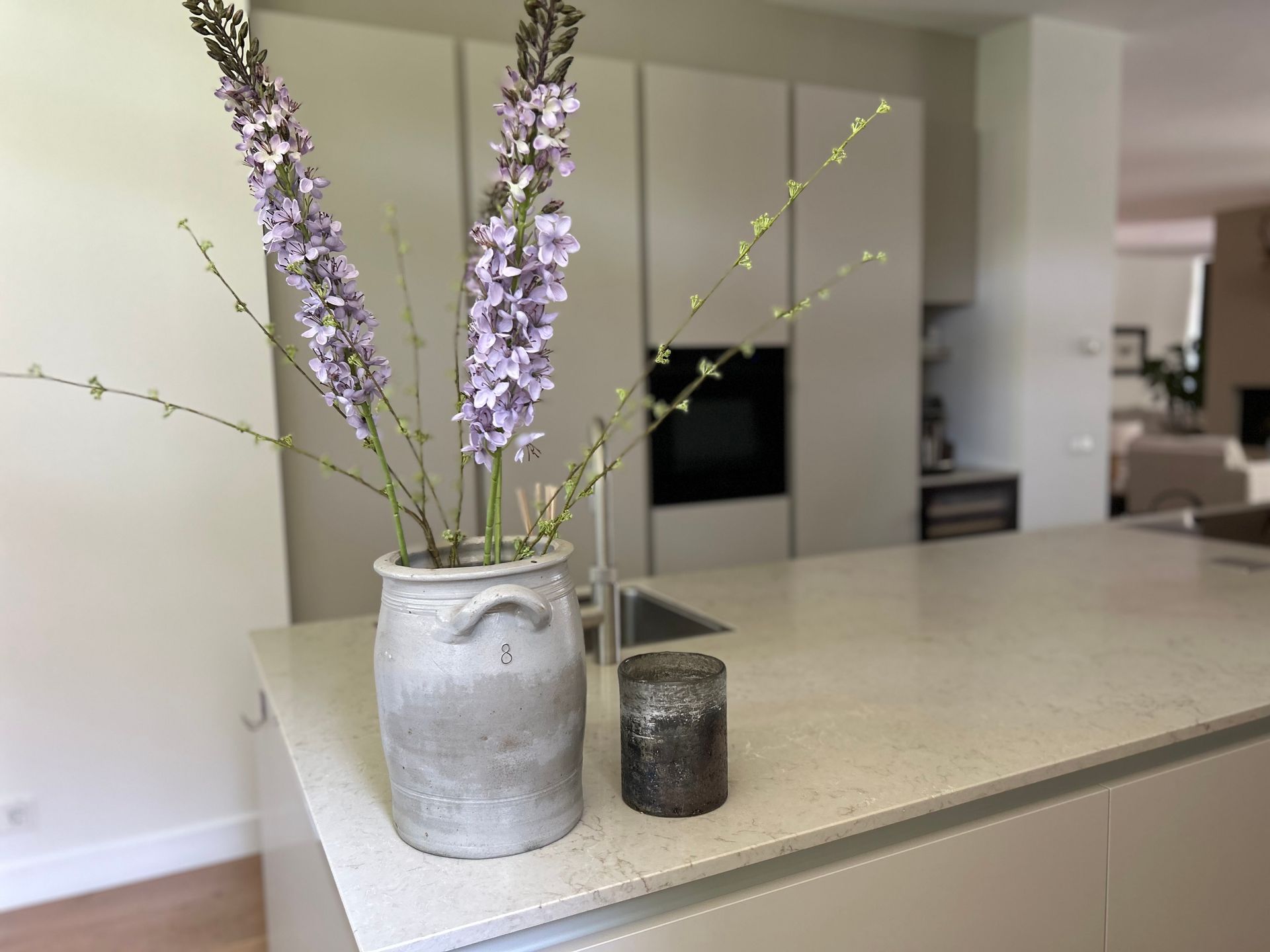 Een vaas met paarse bloemen op een keukeneiland naast een klein kaarsje, met een moderne keuken op de achtergrond.