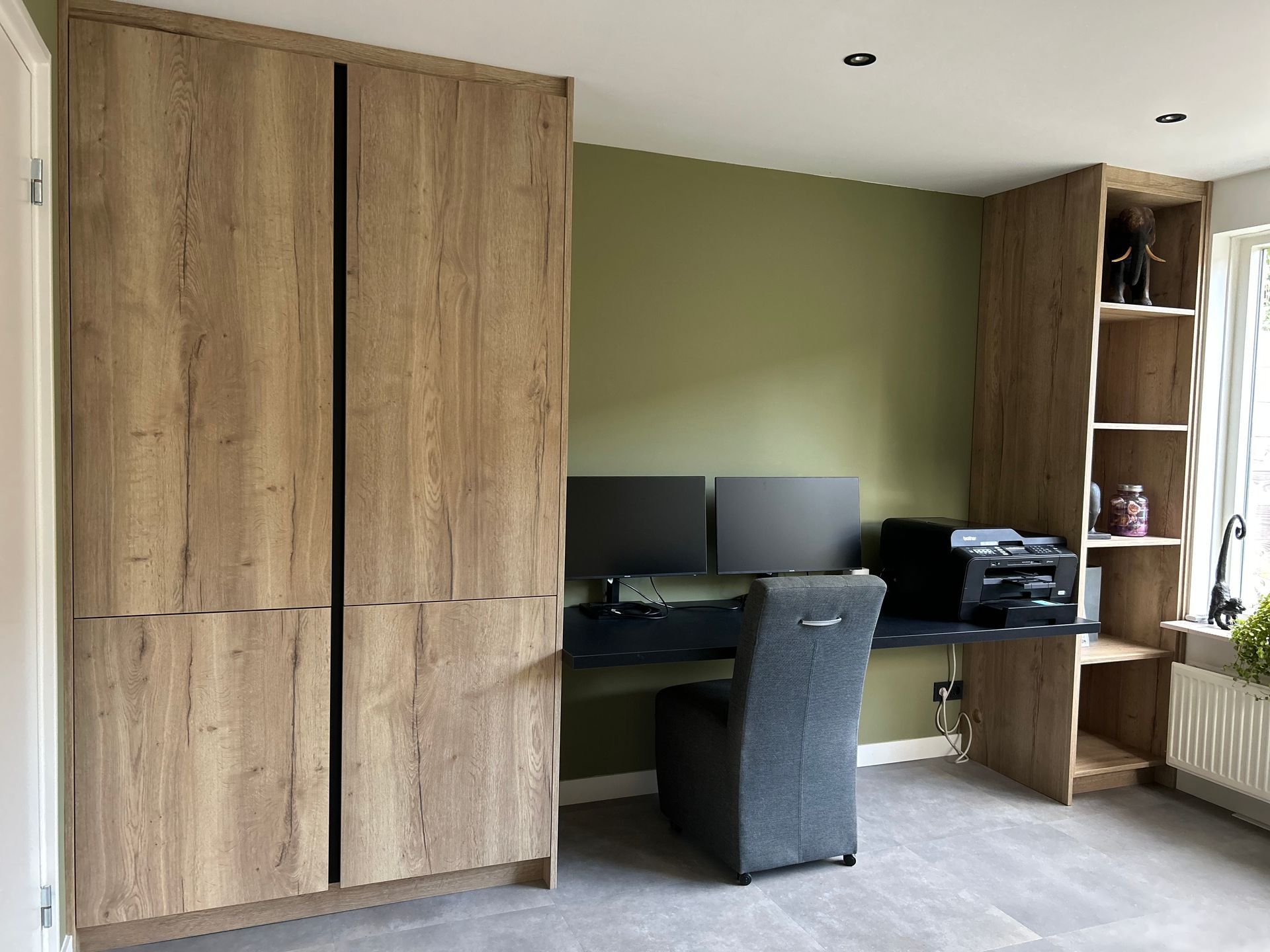 Thuiskantoor met houten kasten, bureau, computermonitor en stoel tegen een olijfgroene muur.