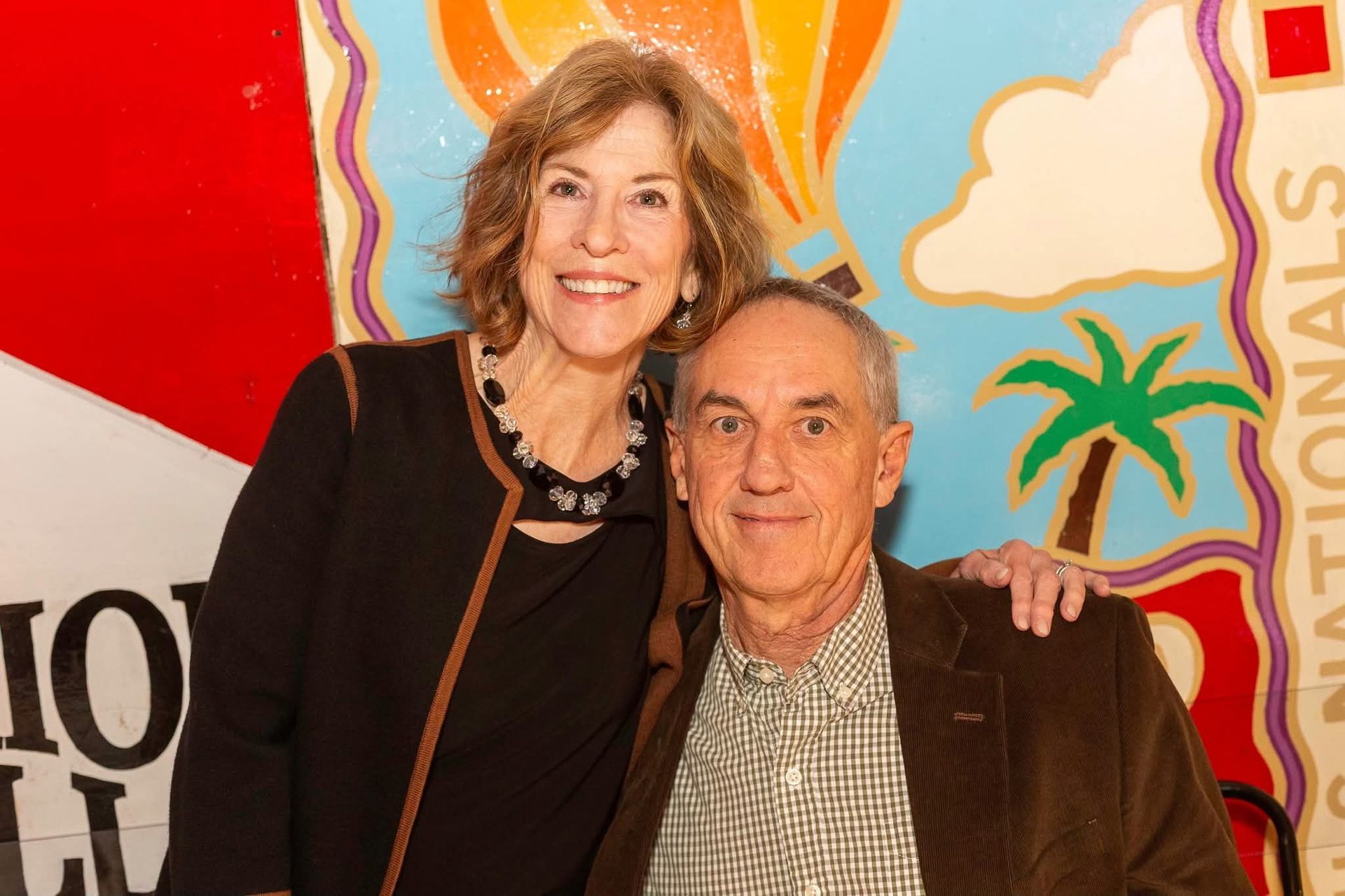 Woman and man smiling, posing in front of a colorful mural. The man has his arm around the woman.