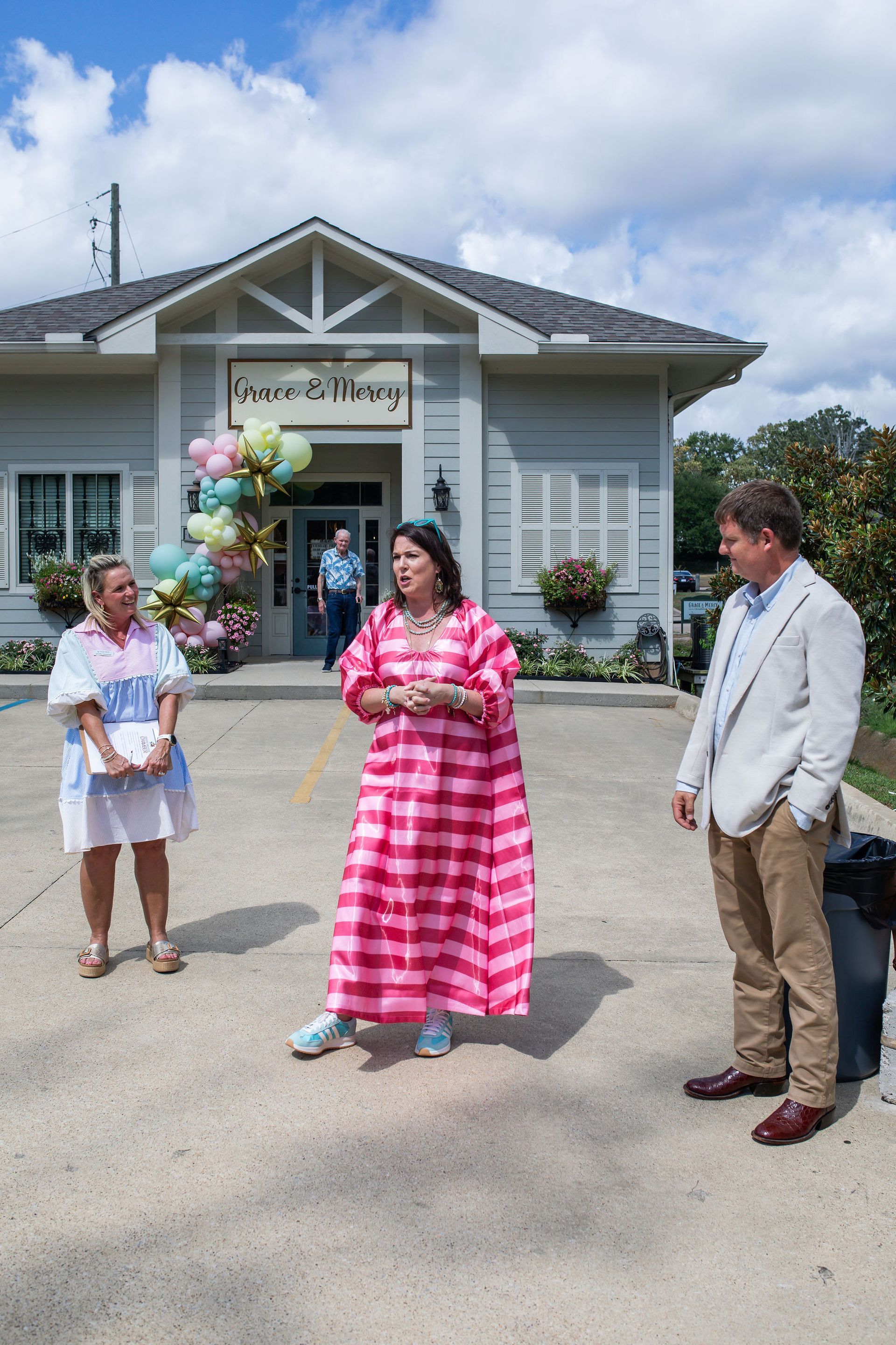 Three people outside a store with balloons. A woman in pink, a woman in white, and a man in khaki.