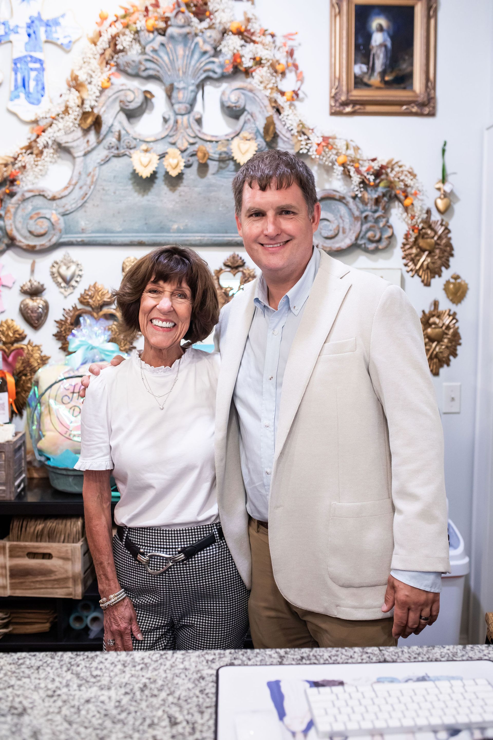 Two people pose inside a shop. Woman wears a white shirt and patterned skirt. Man wears a blazer. Shop decor behind them.