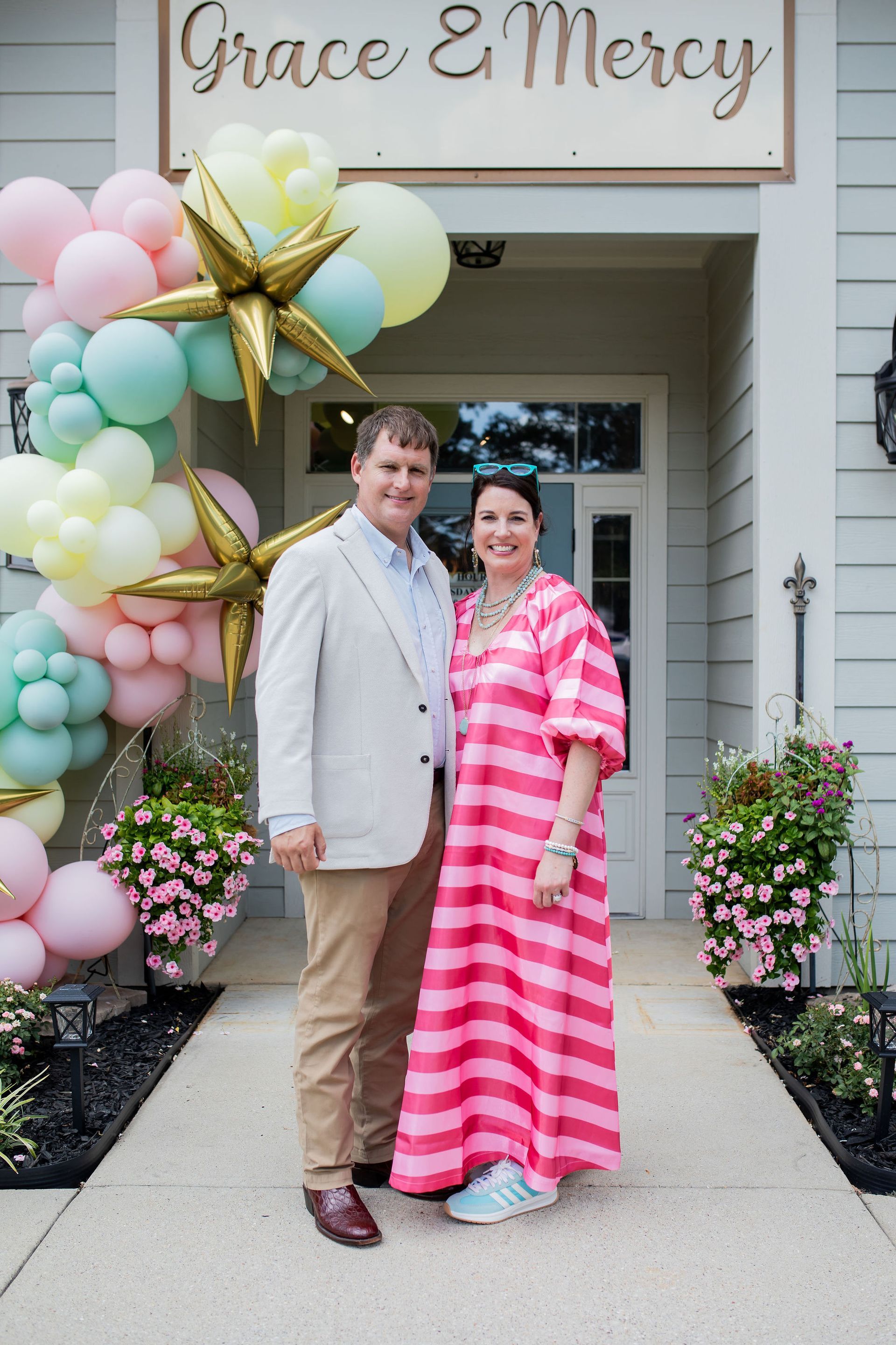 Couple stands outside a shop entrance, under balloon arch. Woman wears pink striped dress. Shop sign reads 