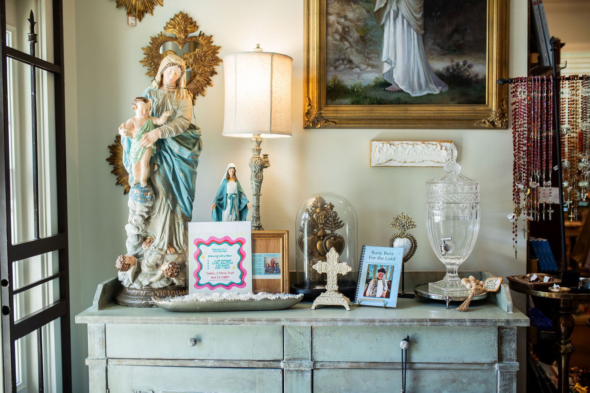 A cabinet displays religious figurines, framed art, cards, a lamp, and glass containers.