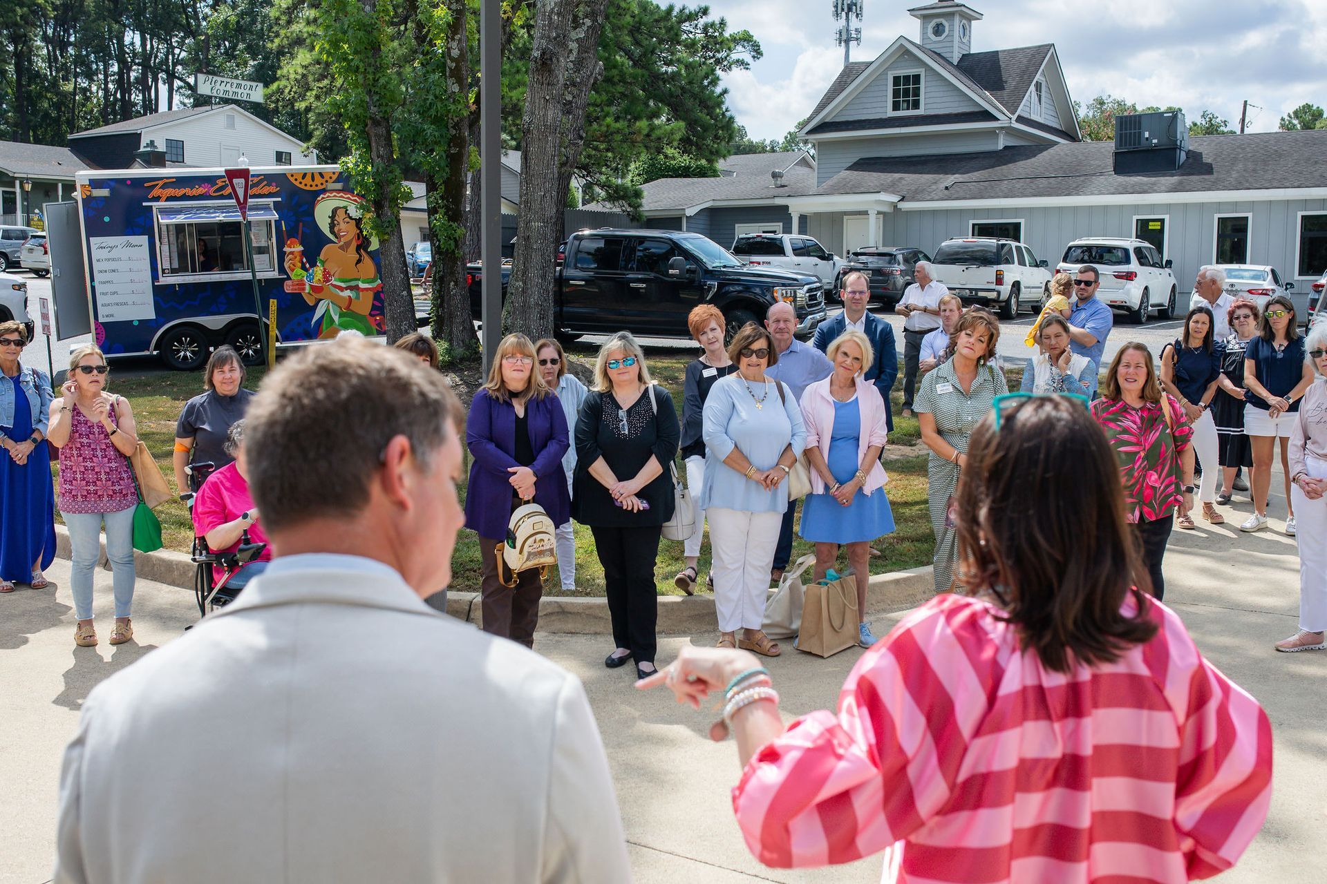 Group of people gathered outdoors, listening to two individuals speaking. Food truck and buildings in background.