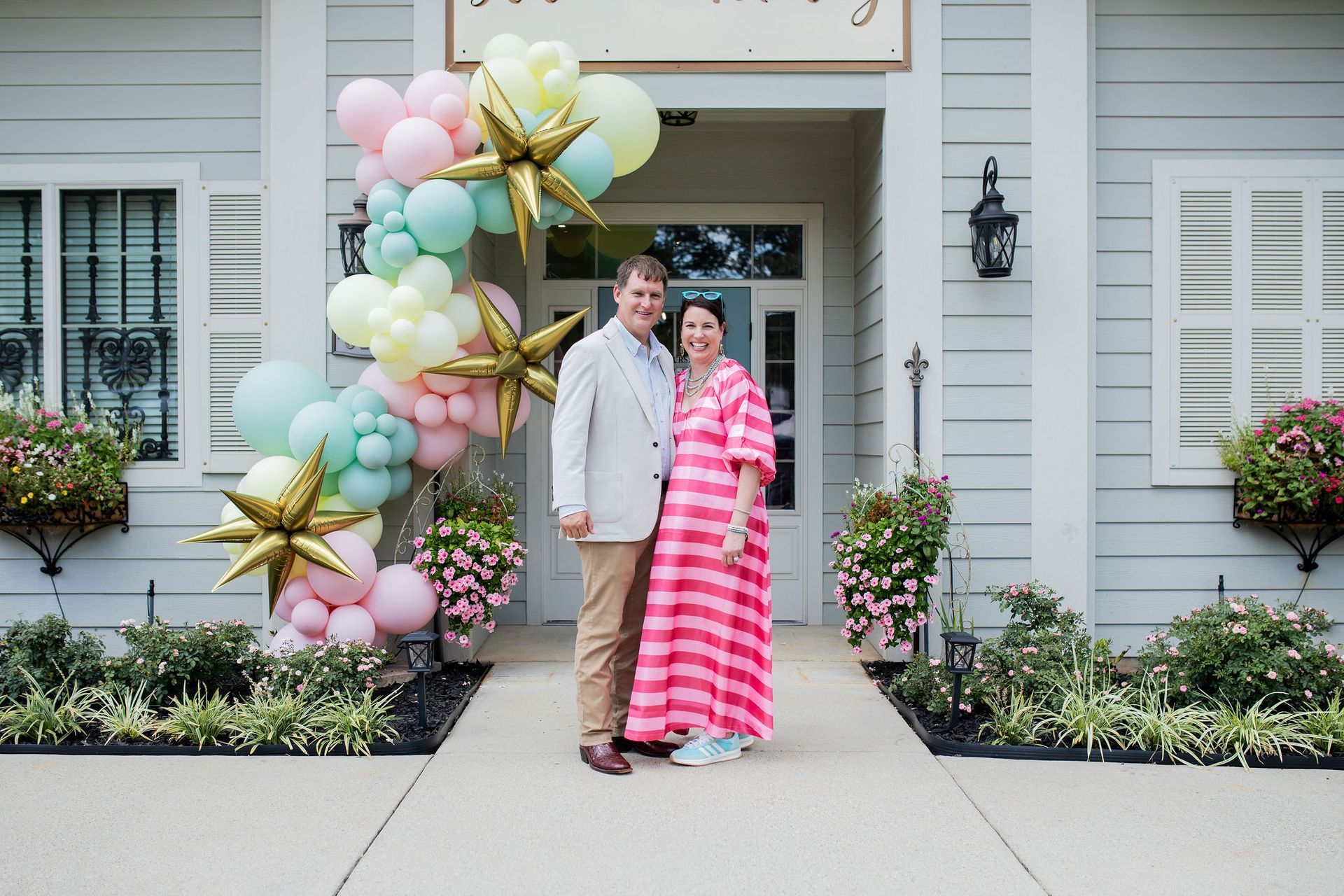 Couple standing in front of a light blue building, adorned with balloons, flowers, and a sign.