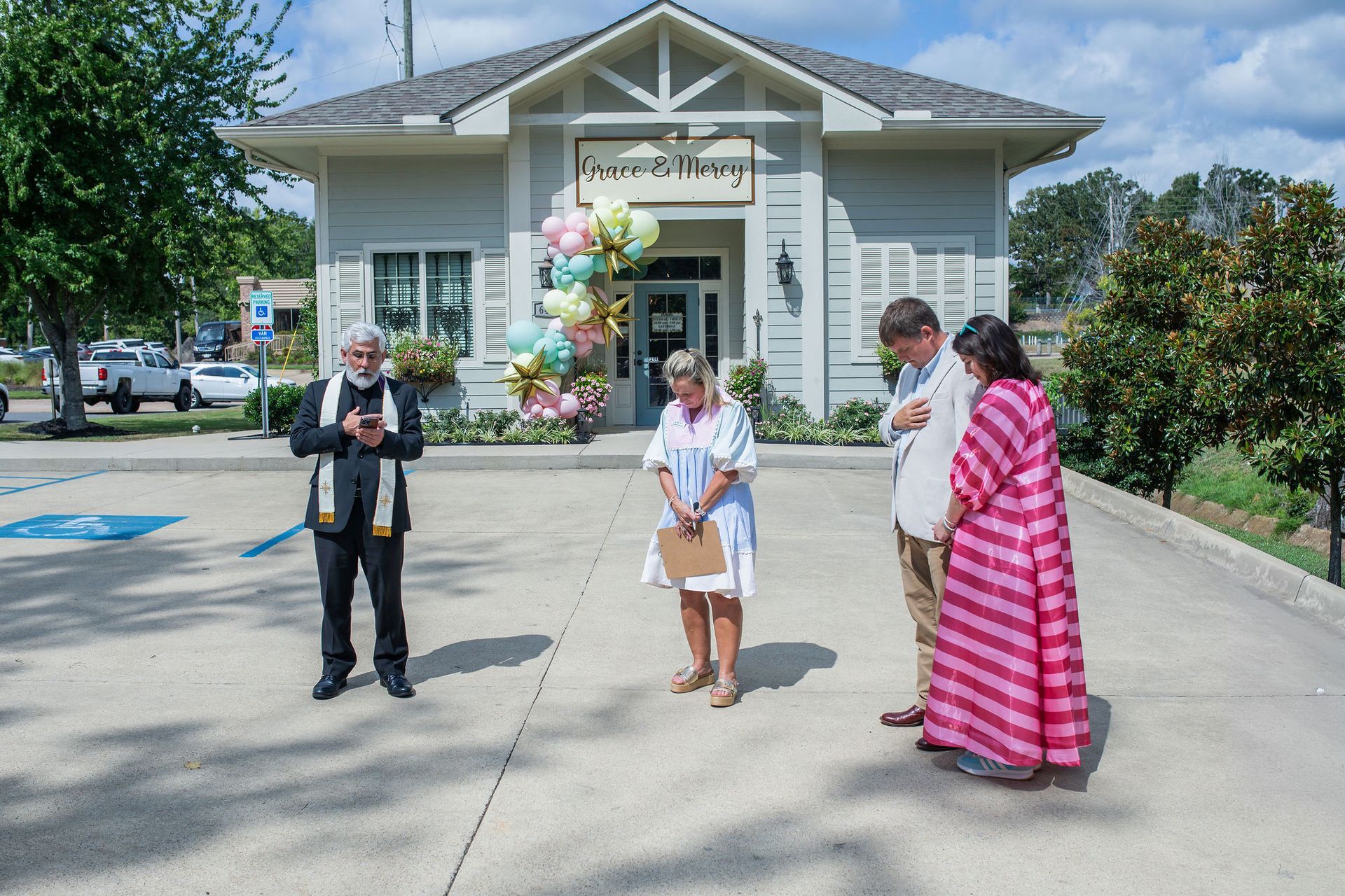A priest and three people stand in front of a building with balloons, possibly a business opening.