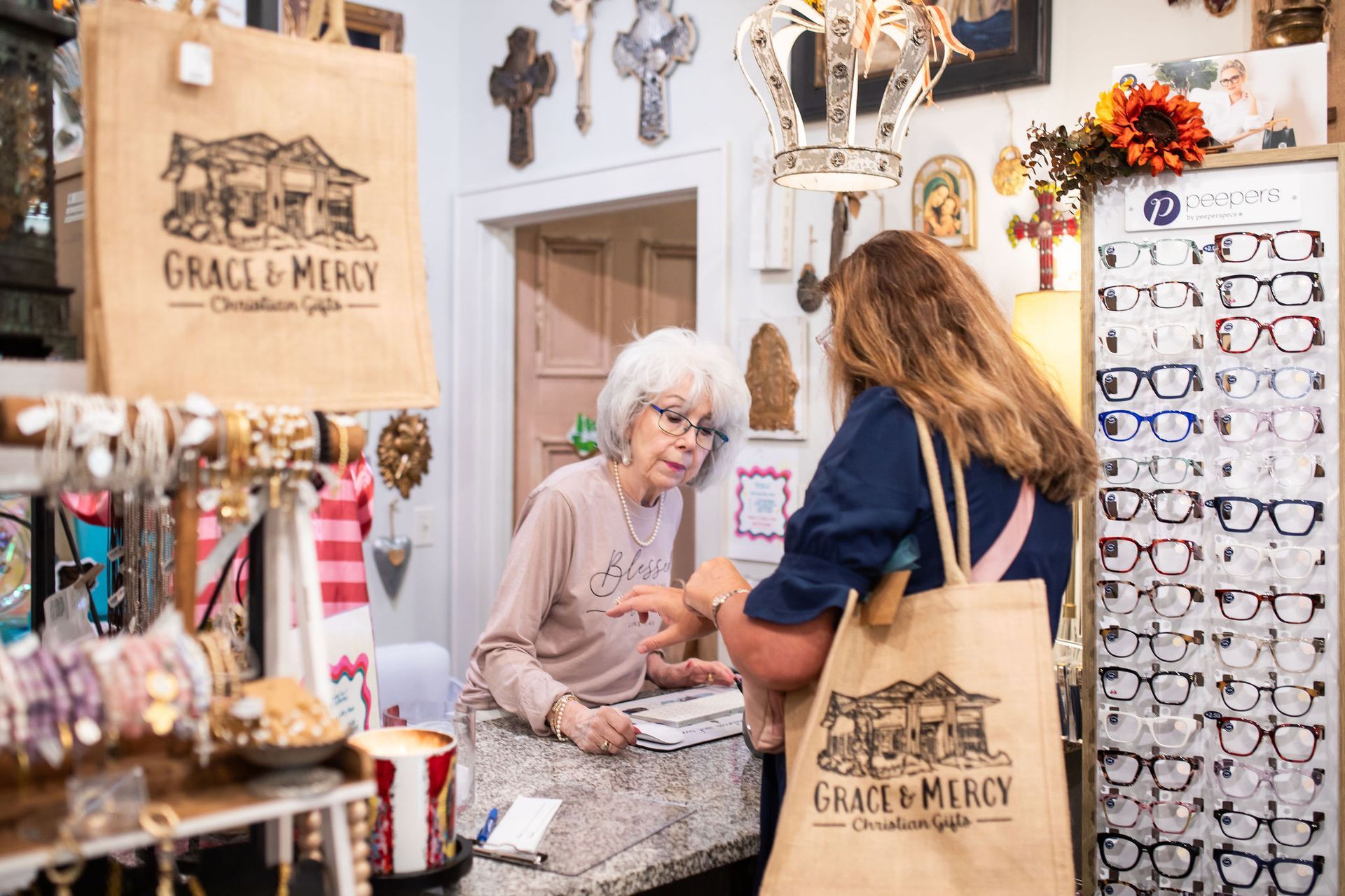 Woman assisting a customer at a shop counter. Both women have shopping bags. Shop interior with religious items and eyeglasses.