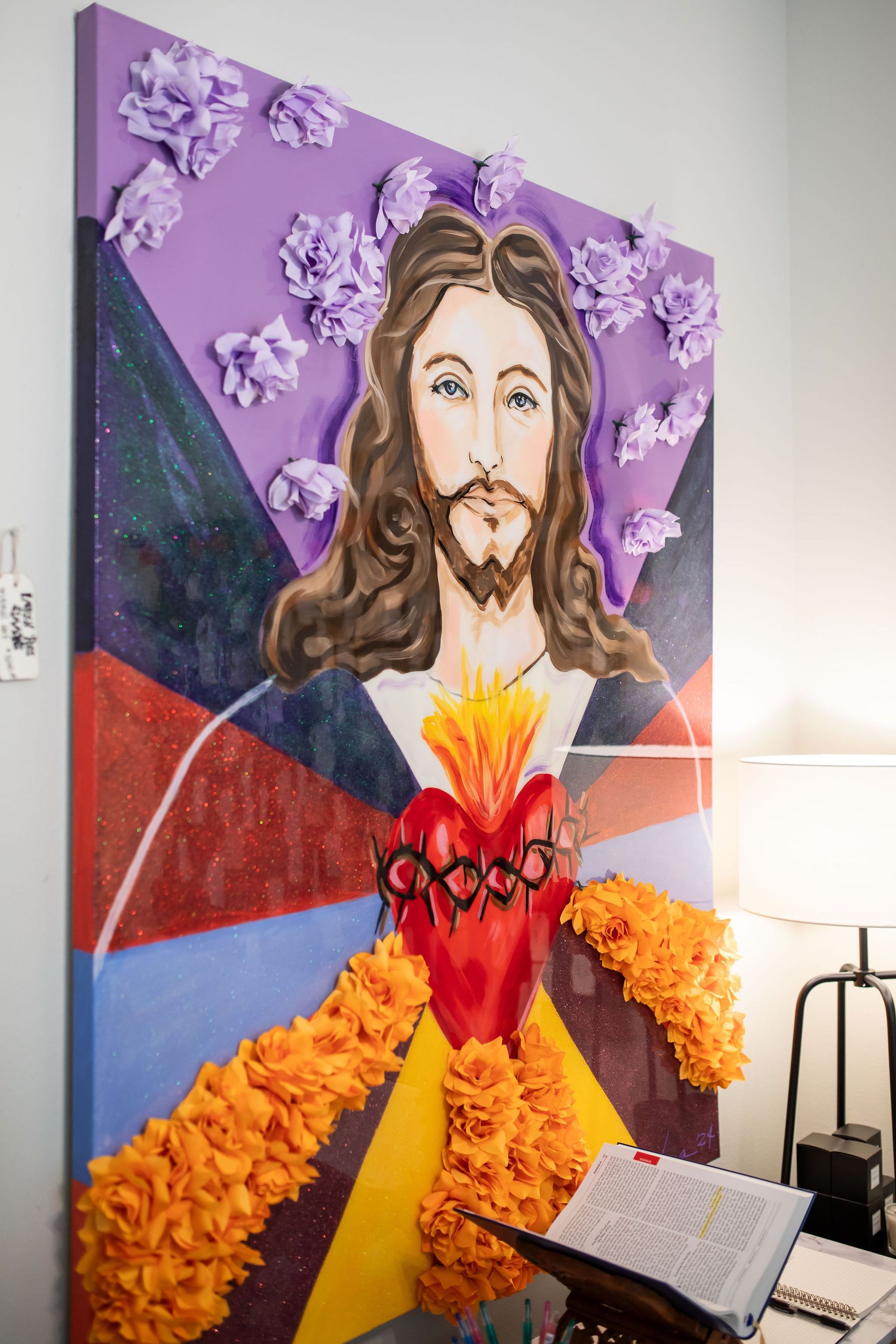 Painting of Jesus with flaming heart, surrounded by floral decorations, against geometric background.