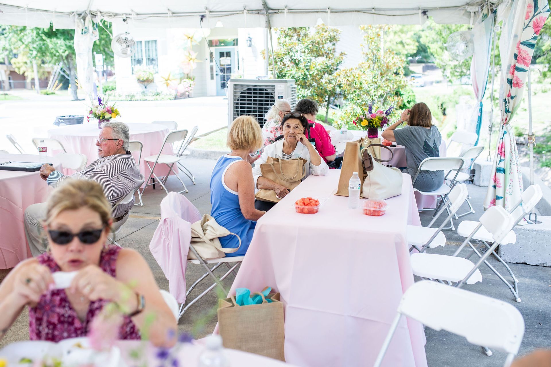 People sitting at tables under a tent; some eating, talking; pale pink tablecloths.